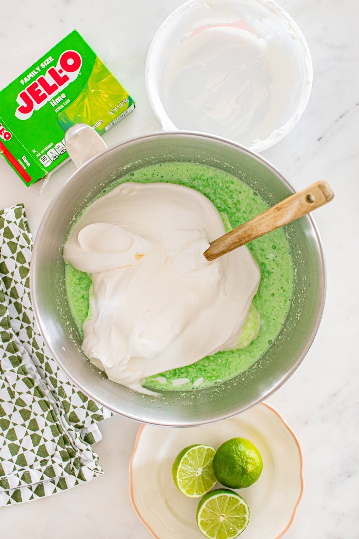 Mixing bowl with green lime Jello and whipped topping, wooden spoon, limes, and Jello box on a countertop.