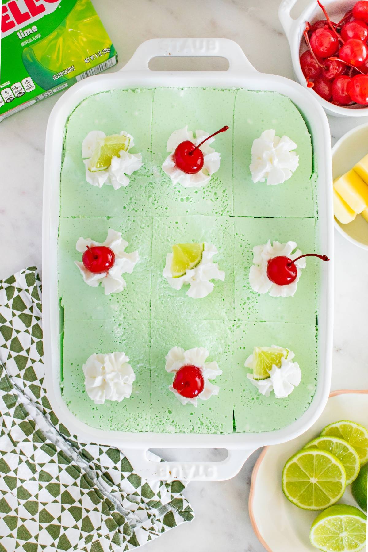 A green Jello salad dessert in a white dish, topped with whipped cream, cherries, and lime slices.