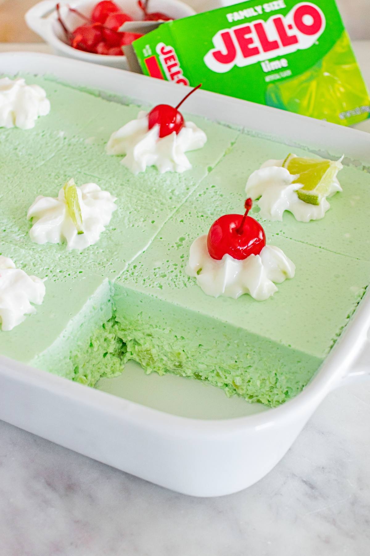 A dish of green Jello salad dessert topped with whipped cream, cherries, and lime slices, with a piece missing.