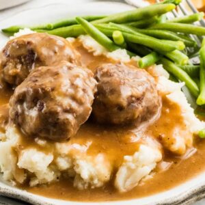 Three meatballs and gravy over mashed potatoes, served with green beans on a white plate.