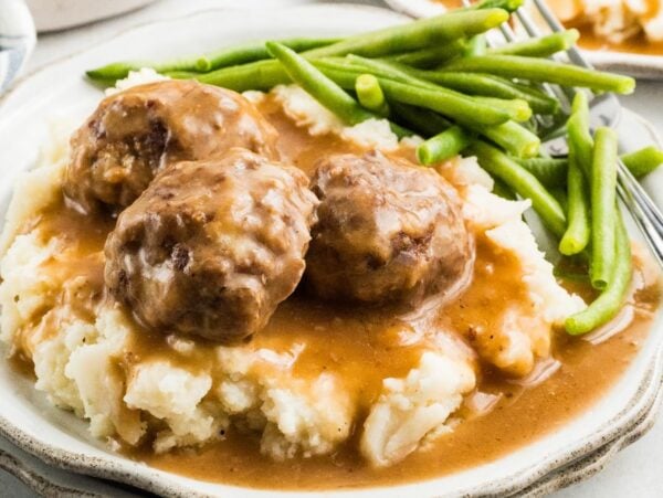 Three meatballs and gravy over mashed potatoes, served with green beans on a white plate.