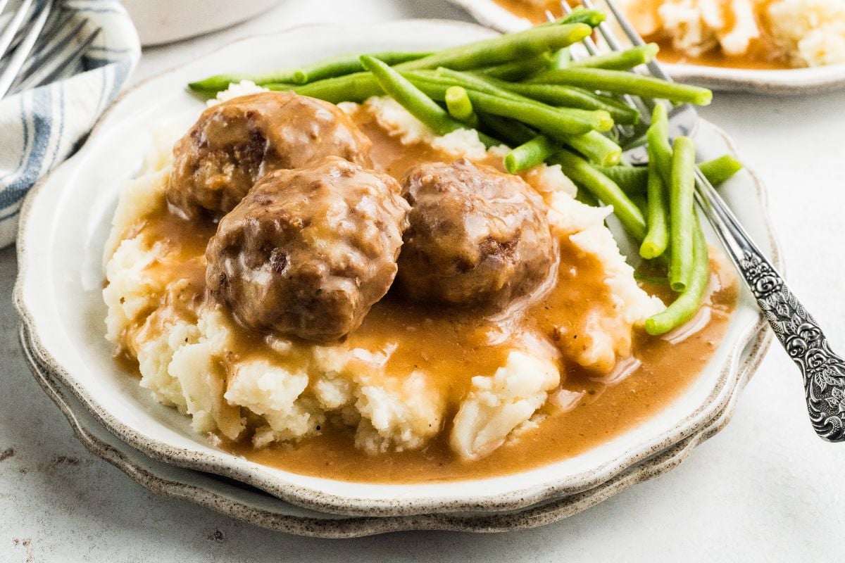 Plate of mashed potatoes topped with meatballs and gravy, served alongside crisp green beans, with a fork on the side.
