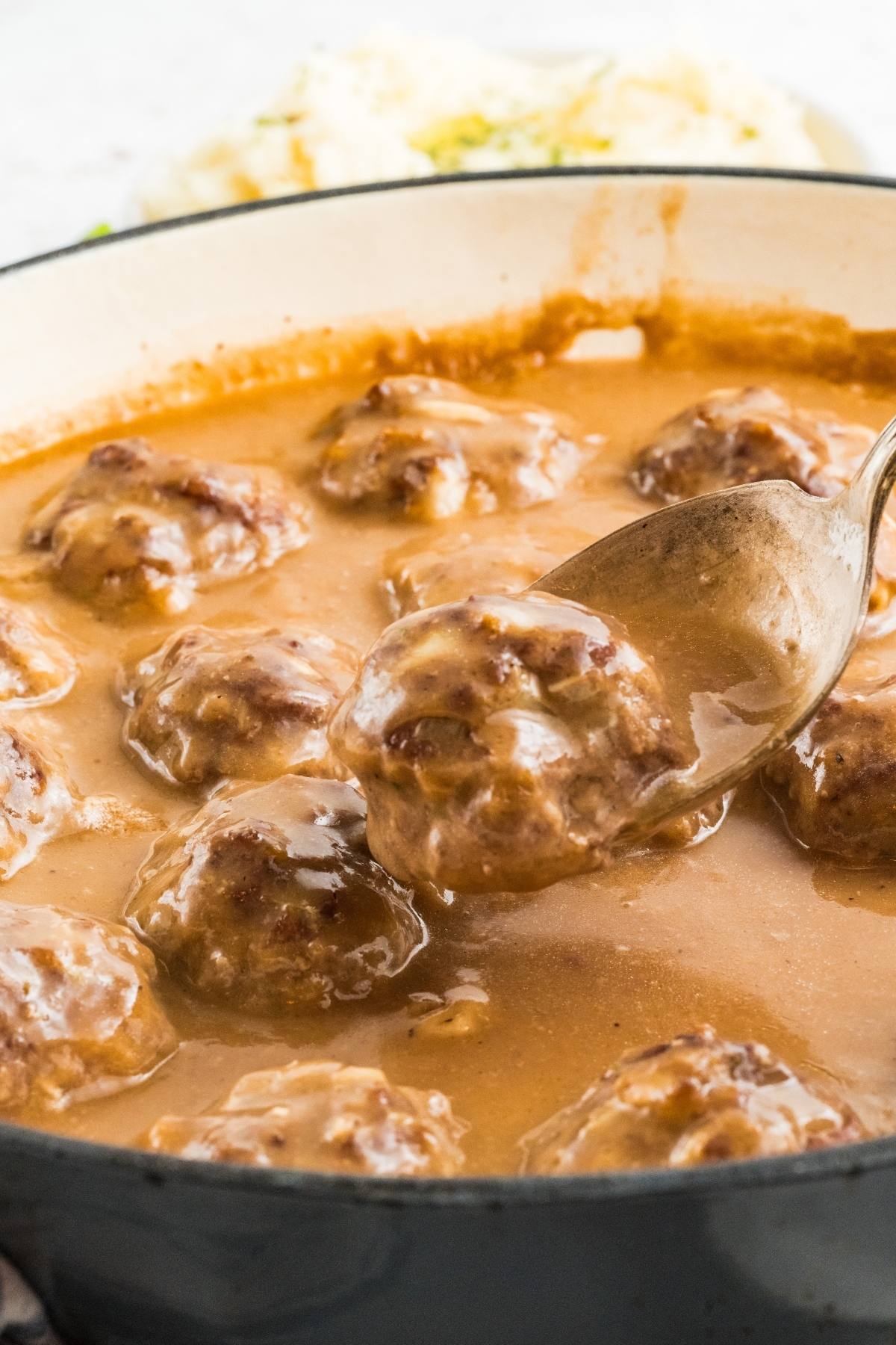 A spoon lifts a meatball covered in brown gravy from a pot filled with meatballs and sauce.