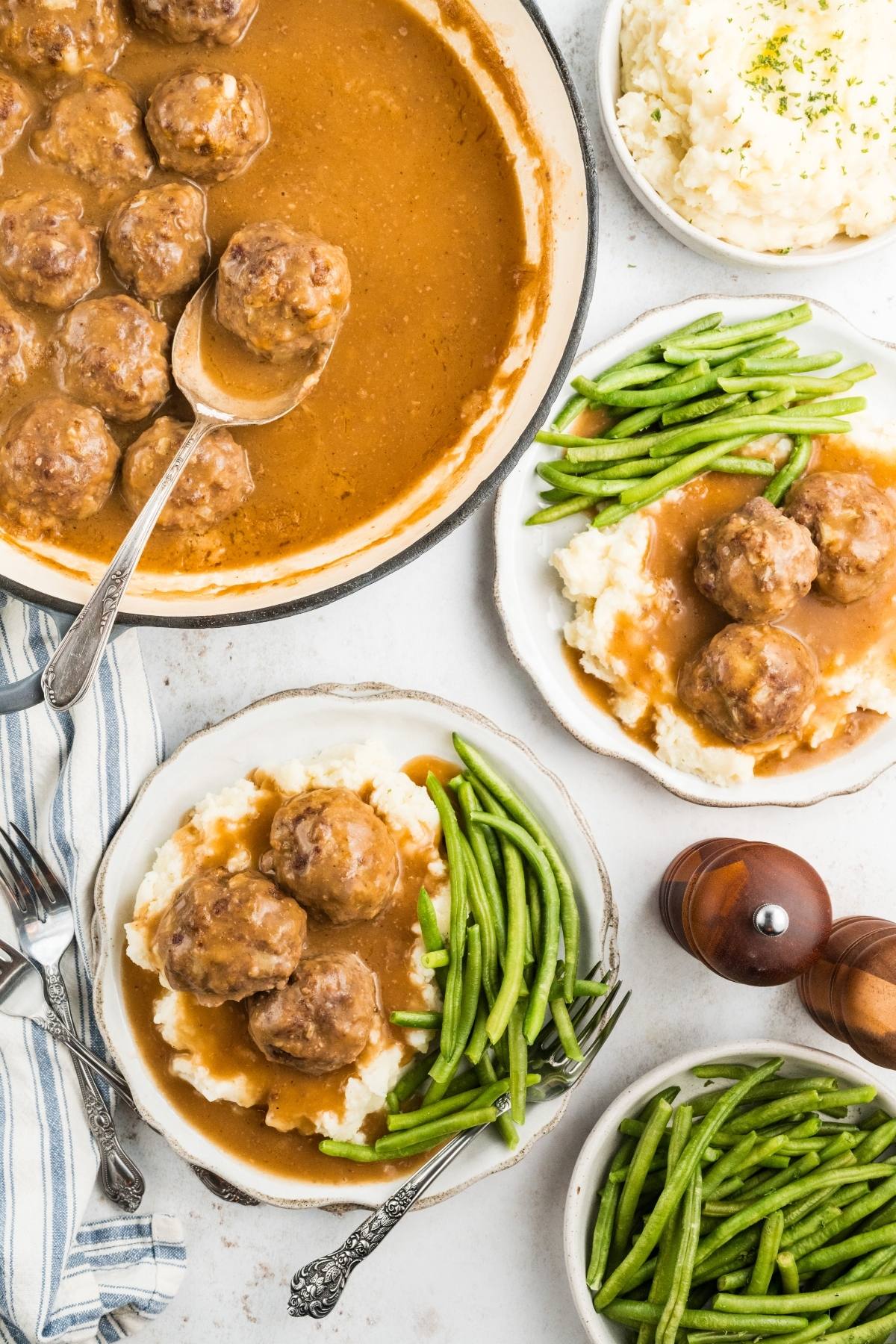 Two plates with meatballs in gravy on mashed potatoes, green beans on the side, plus extra bowls of sides.