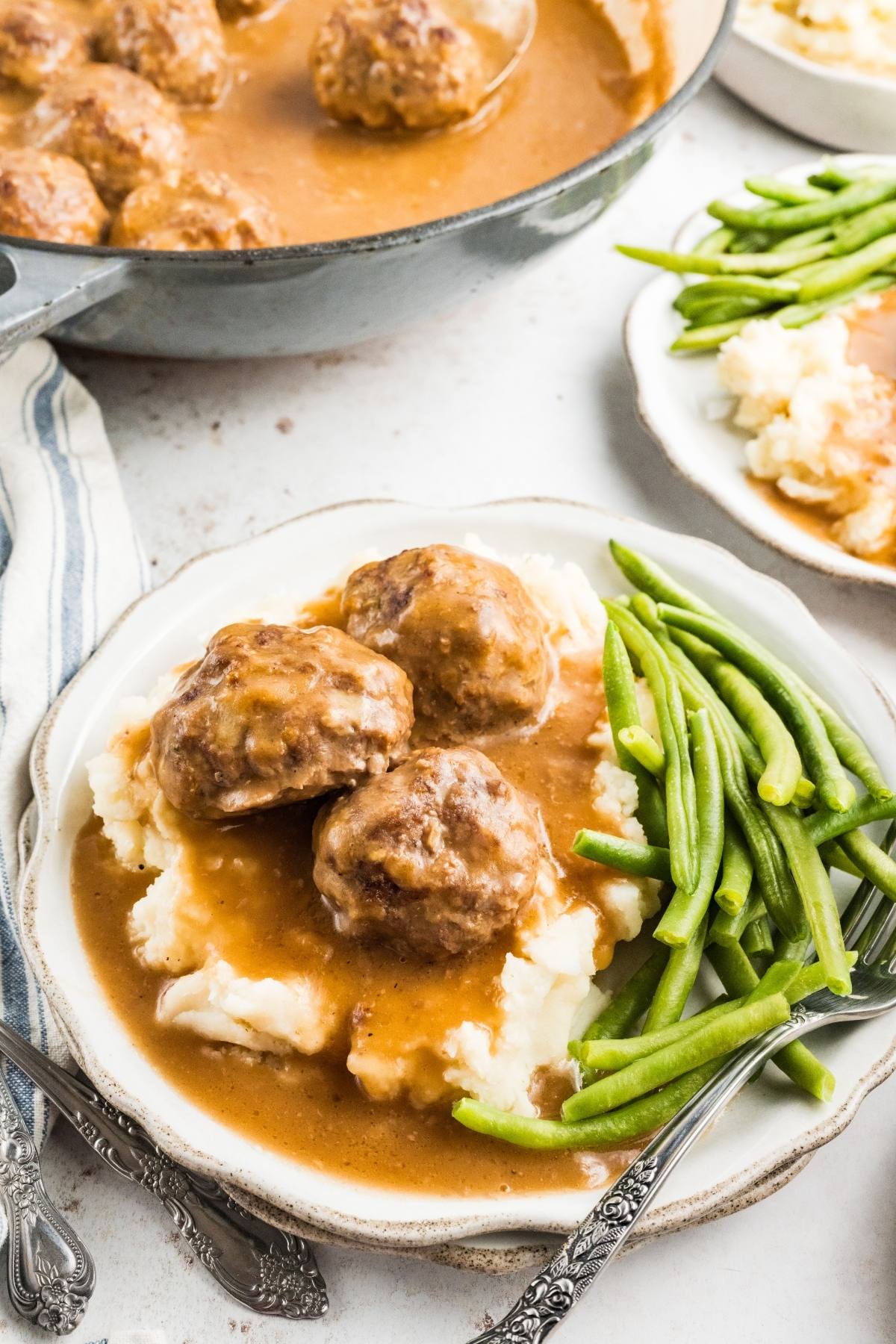 Plate with mashed potatoes, meatballs in brown gravy, and green beans, with a pan of meatballs in the background.