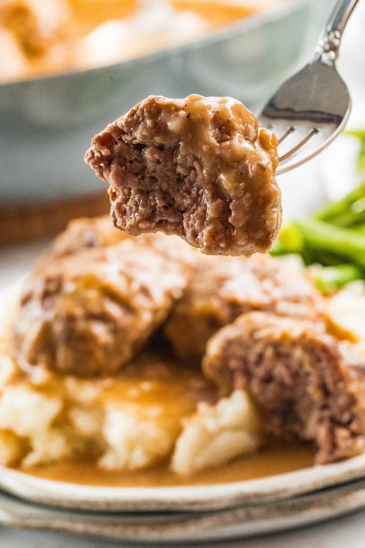 A fork holding a bite of Salisbury steak with gravy, with mashed potatoes and steak on a plate in the background.
