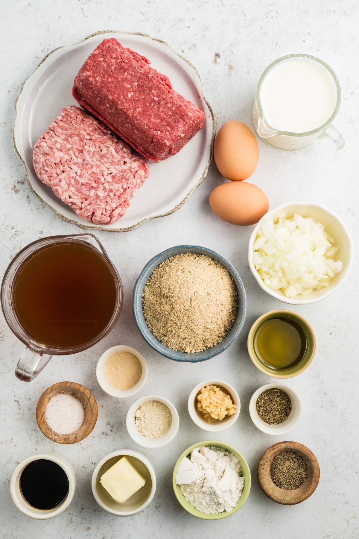 Ground beef, ground pork, eggs, milk, onion, broth, breadcrumbs, and various spices arranged on a white surface.