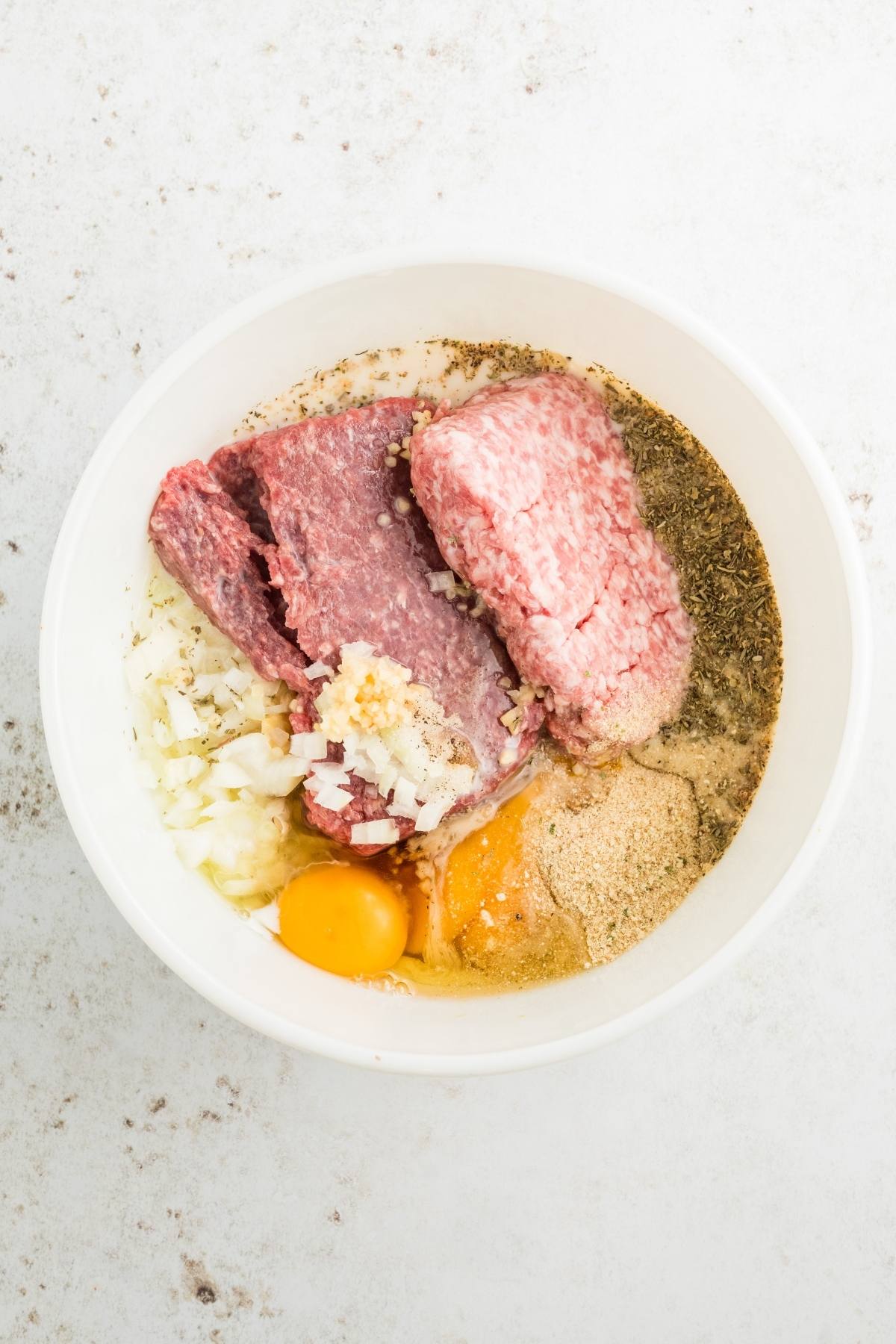 A bowl with ground meat, chopped onions, eggs, breadcrumbs, and seasonings on a white surface.