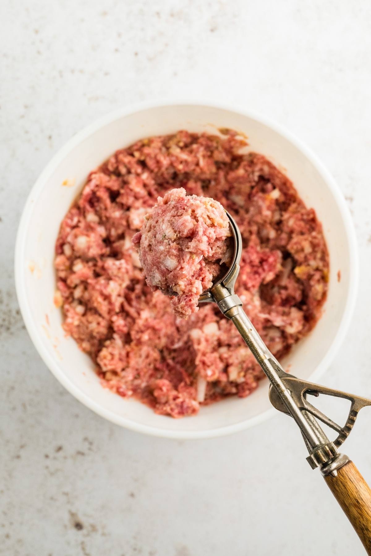 A scoop of raw ground meat mixture held above a white bowl filled with more of the same mixture.