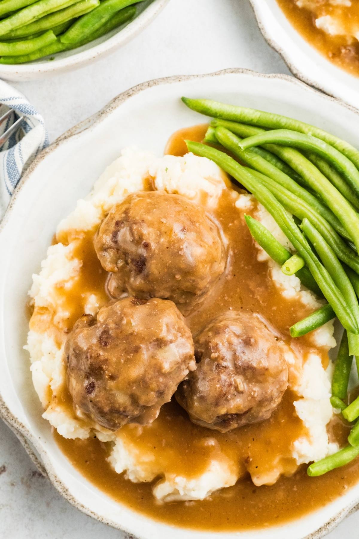 Three meatballs with gravy on mashed potatoes, served with green beans on a white plate.