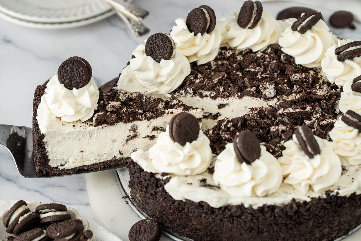 No Bake Oreo Cheesecake topped with whipped cream and mini chocolate sandwich cookies, with a delicious slice being served.