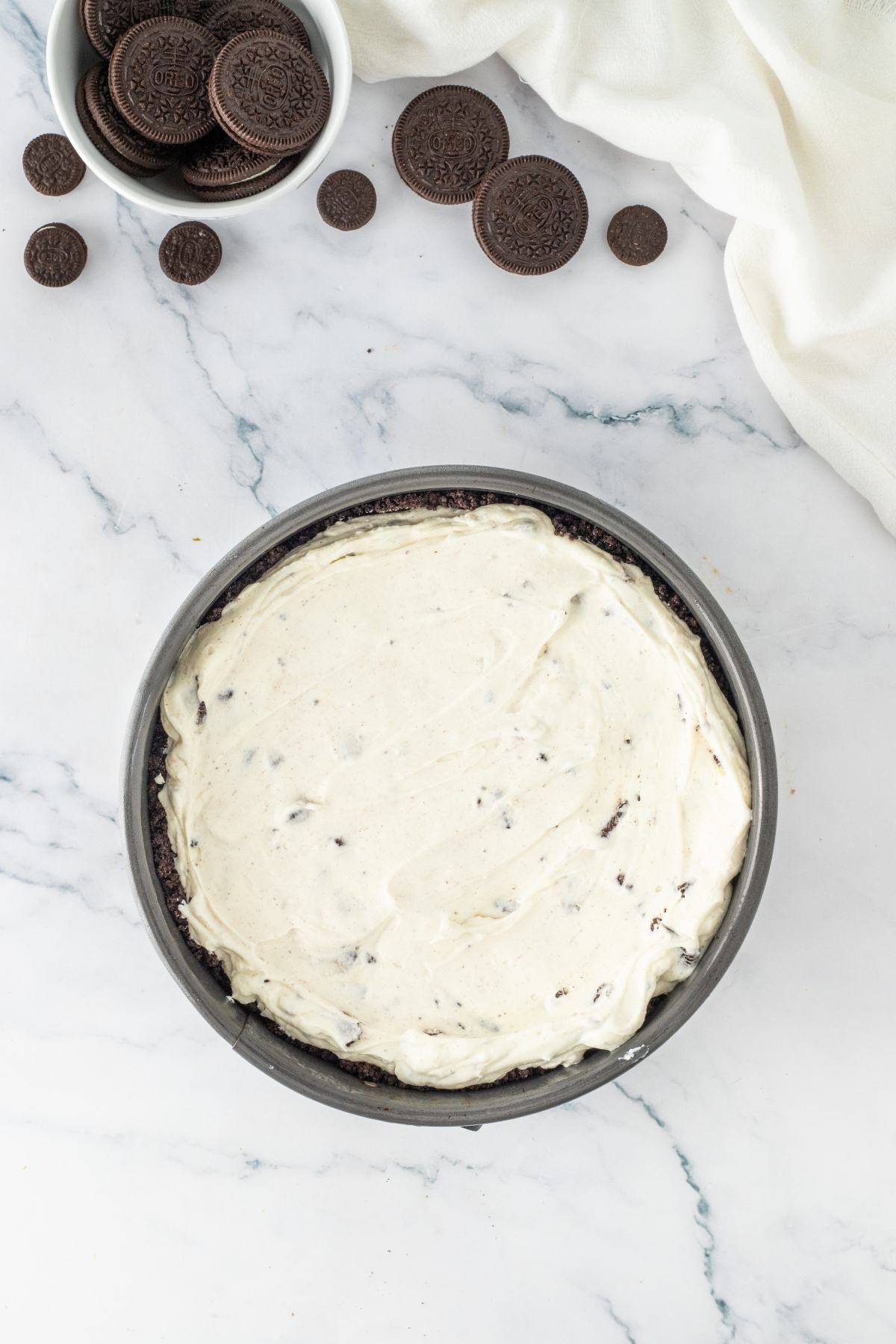 A round pan with Oreo cheesecake filling on a marble surface, with Oreo cookies nearby.