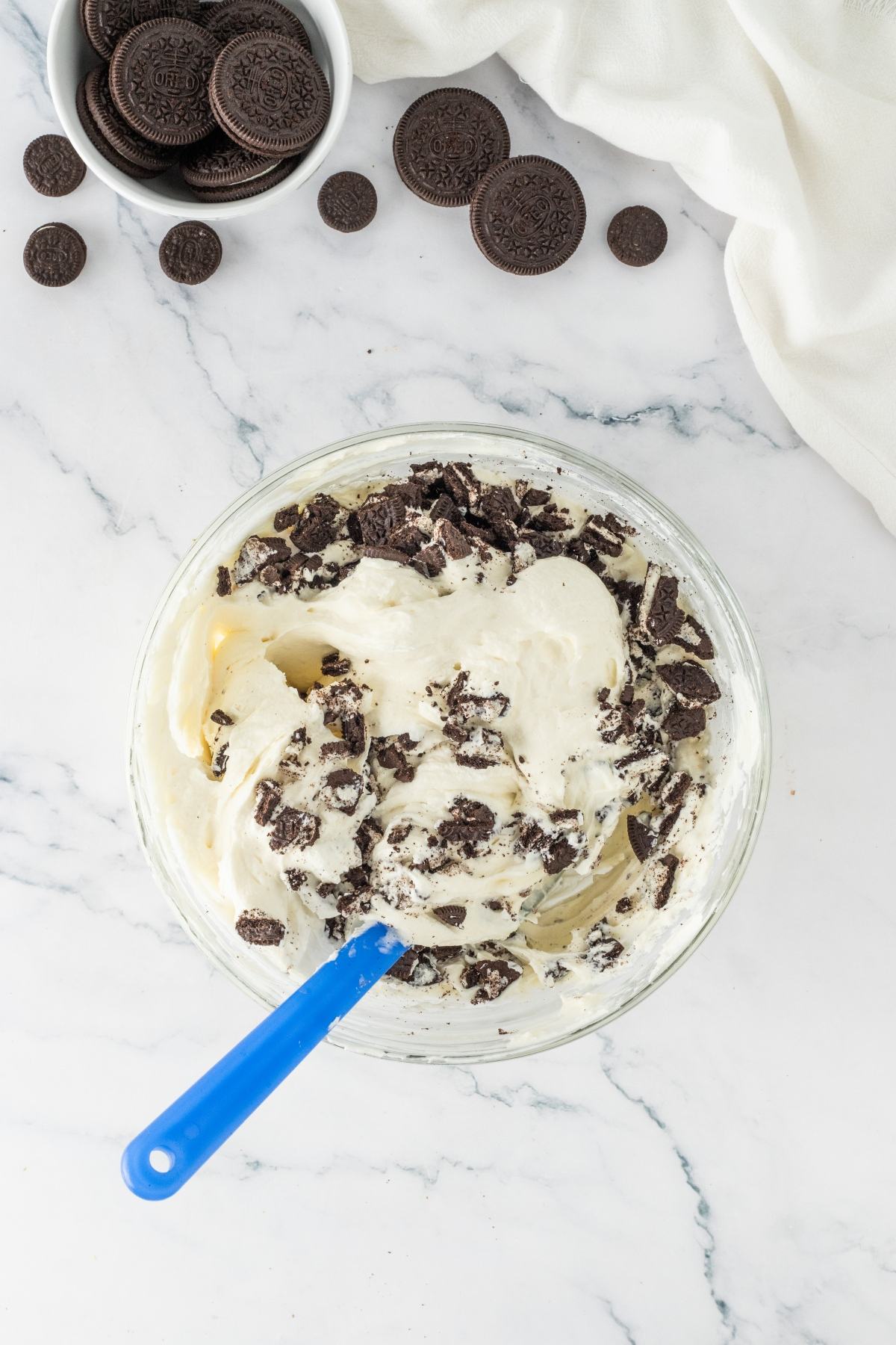 A glass bowl of cream mixed with crushed cookies and a blue spatula, with cookies scattered nearby.