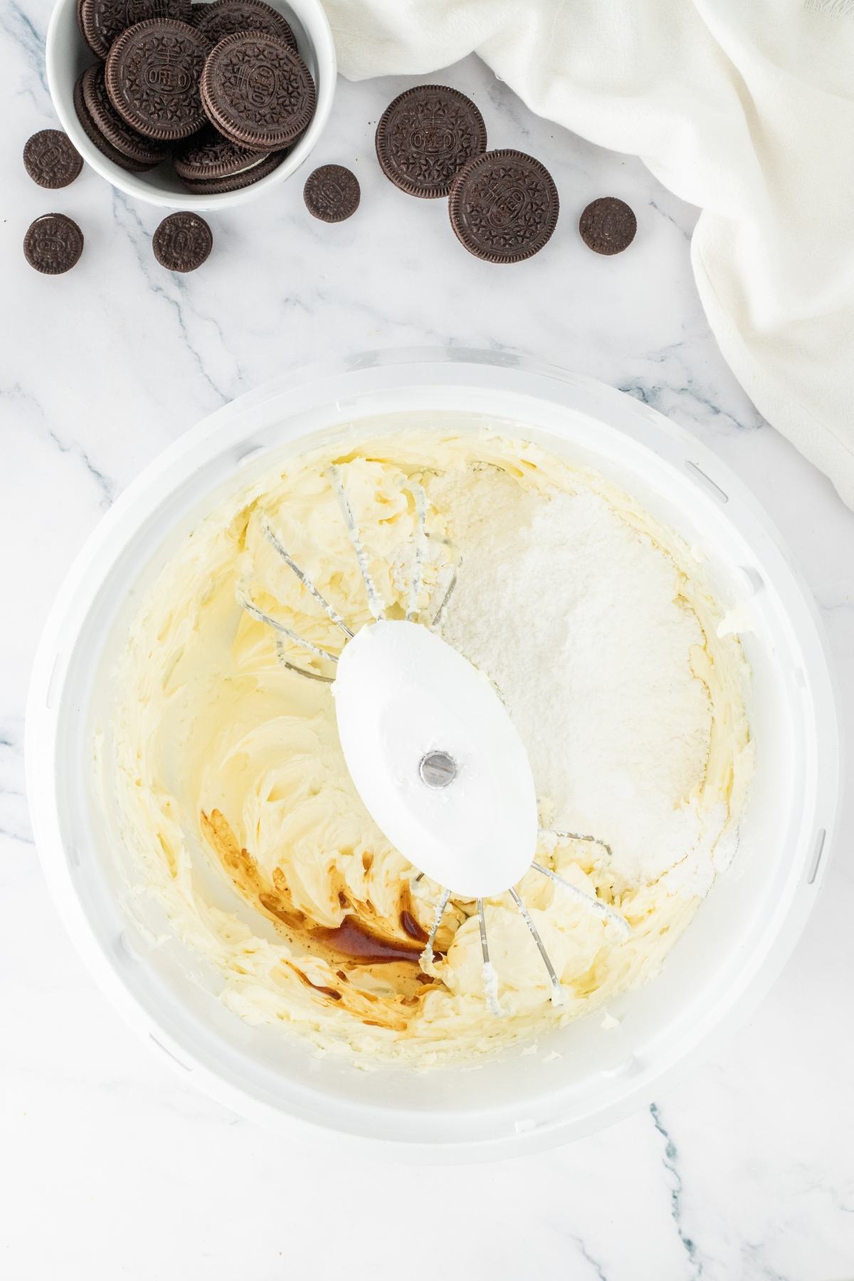 A mixing bowl with cream, vanilla, and flour next to a bowl of chocolate sandwich cookies on a marble surface.