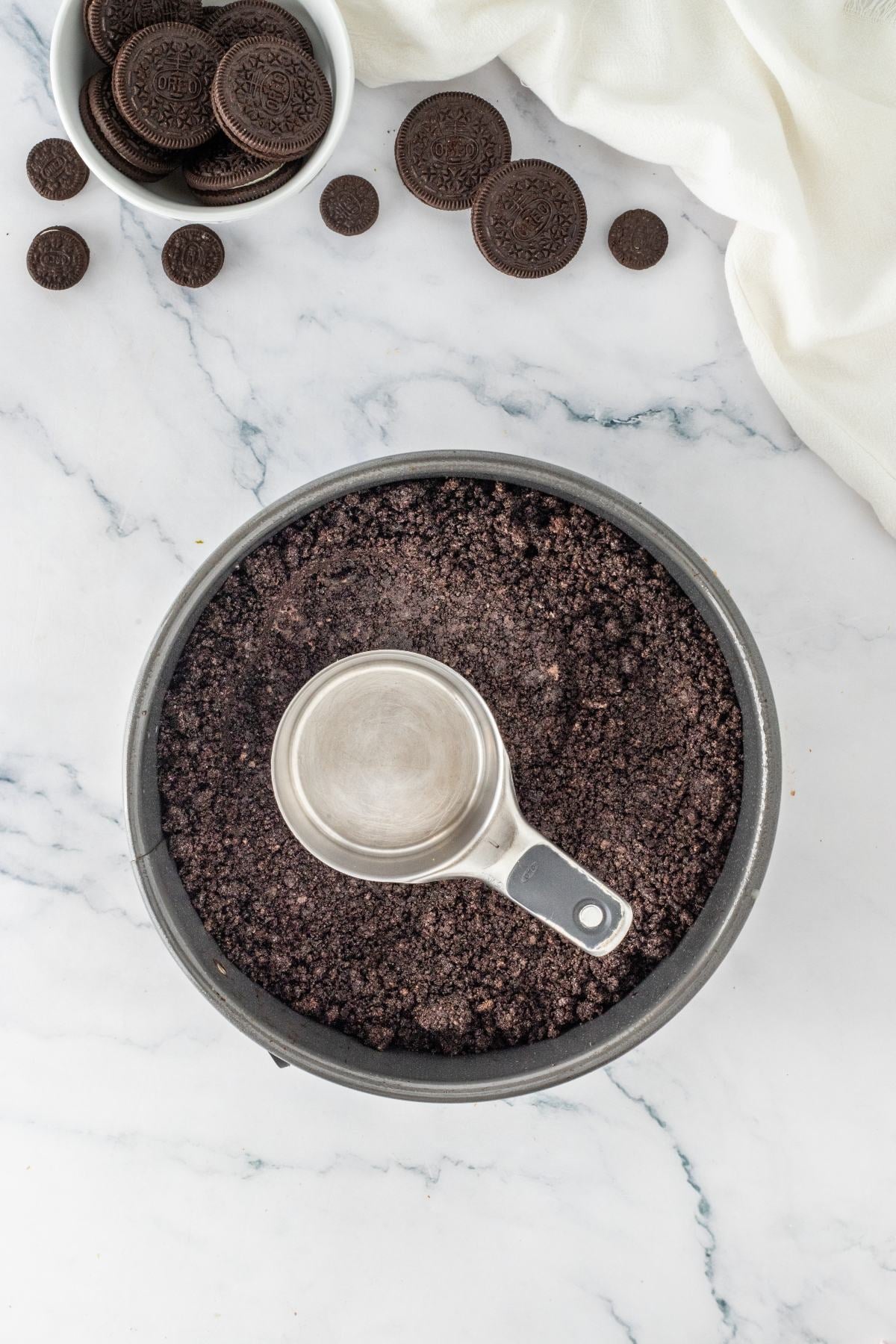 A measuring cup presses Oreo cookie crumbs into a round pan, with whole cookies in a bowl nearby.