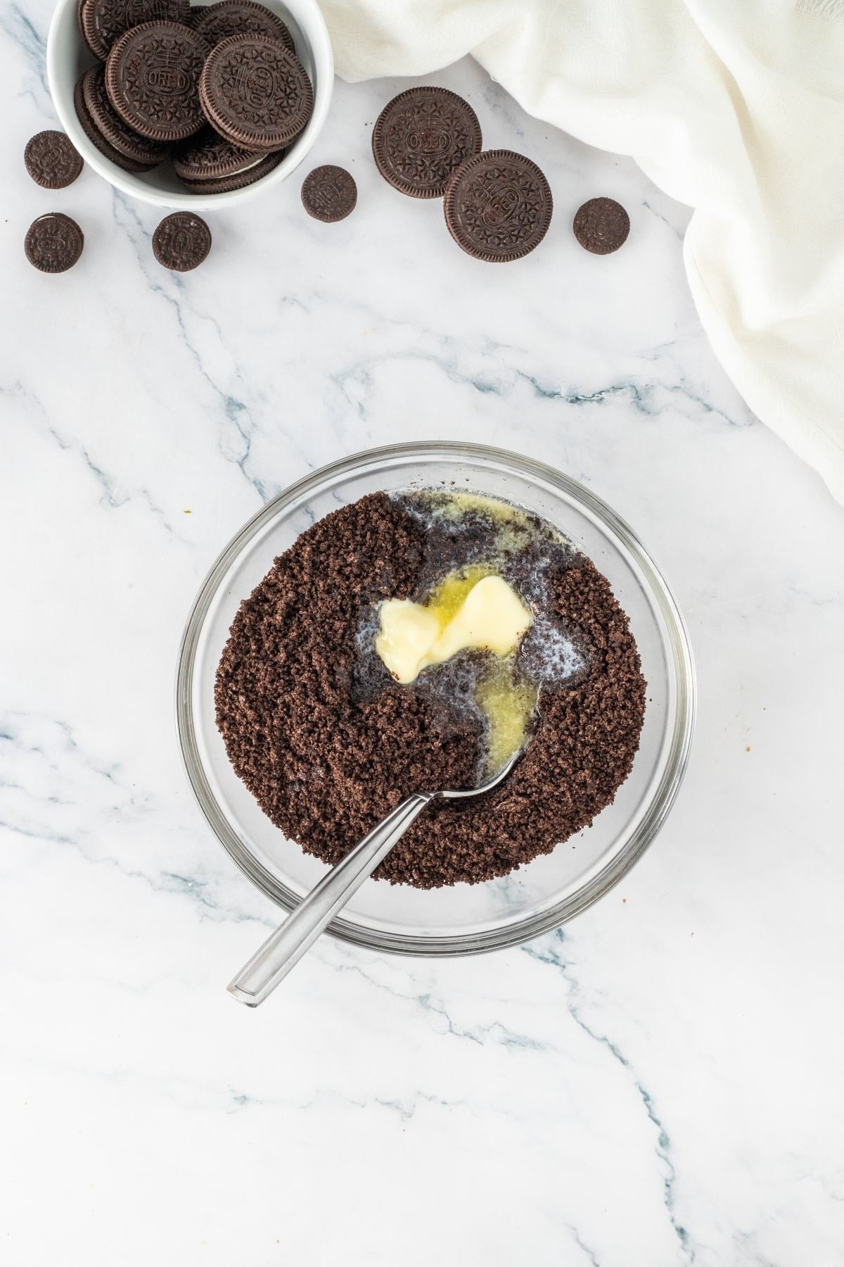 A bowl of crushed cookies and melted butter with a spoon, near whole cookies on a marble surface.