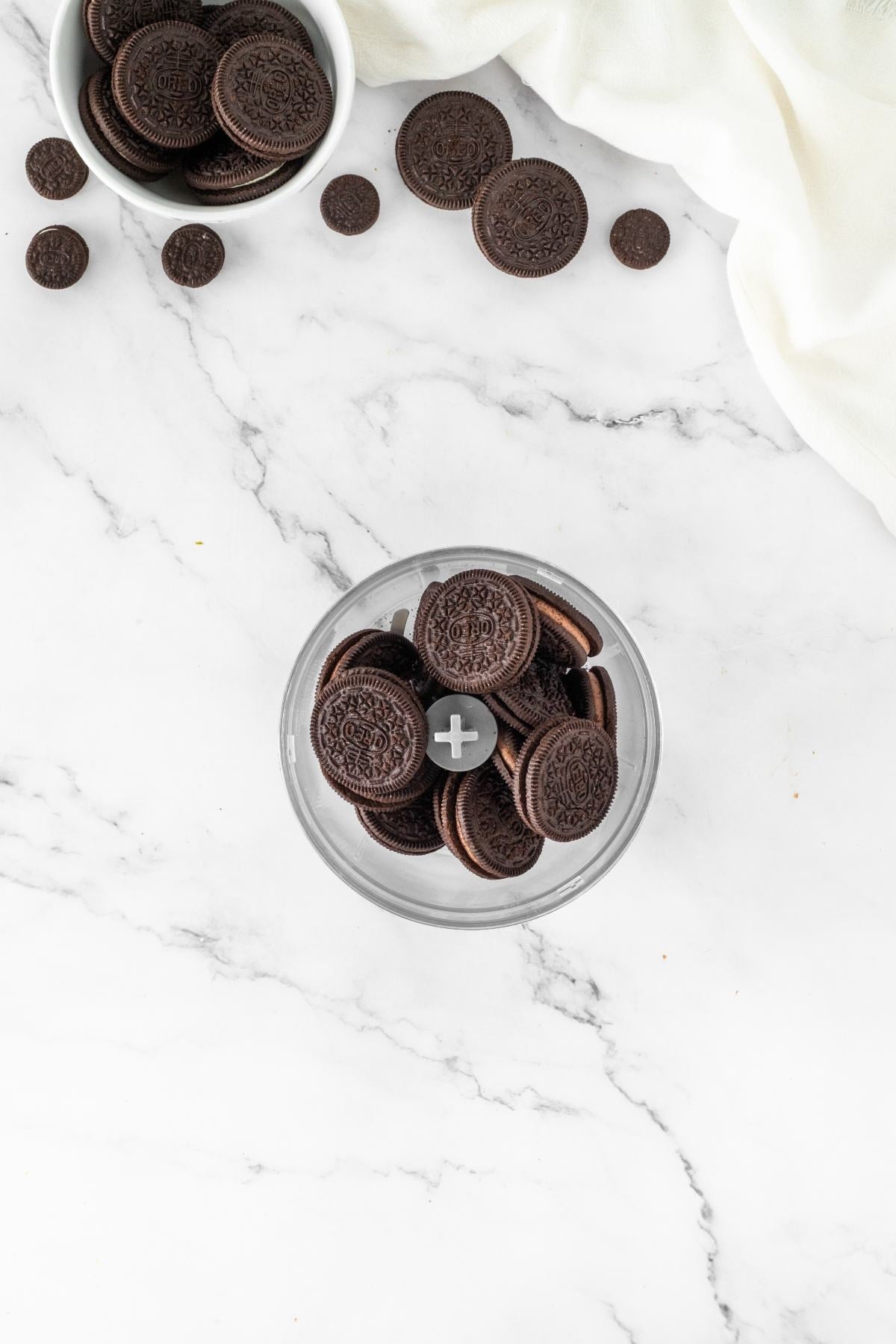 Chocolate sandwich cookies in a food processor on a marble countertop, with cookies scattered nearby.