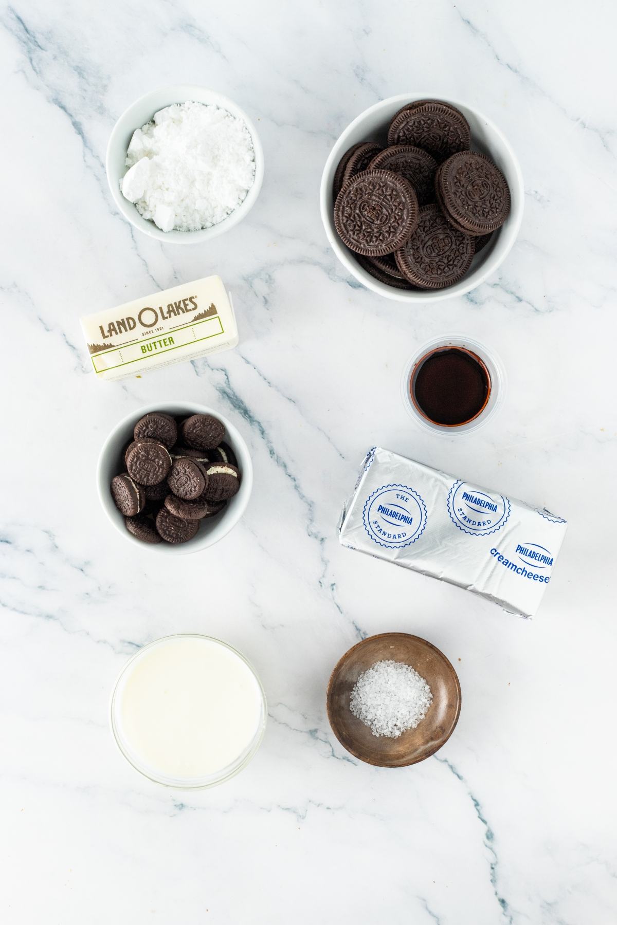 Baking ingredients on a marble surface: cookies, cream cheese, butter, milk, powdered sugar, vanilla, and salt.