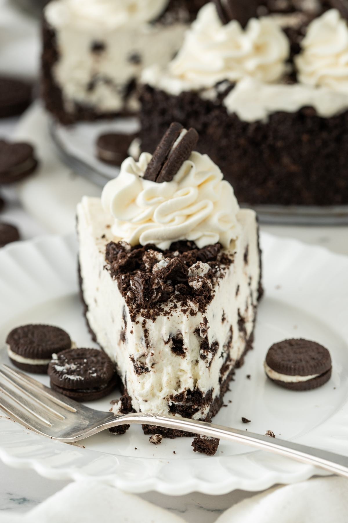A slice of cookies and cream cheesecake topped with whipped cream and mini cookies on a white plate.