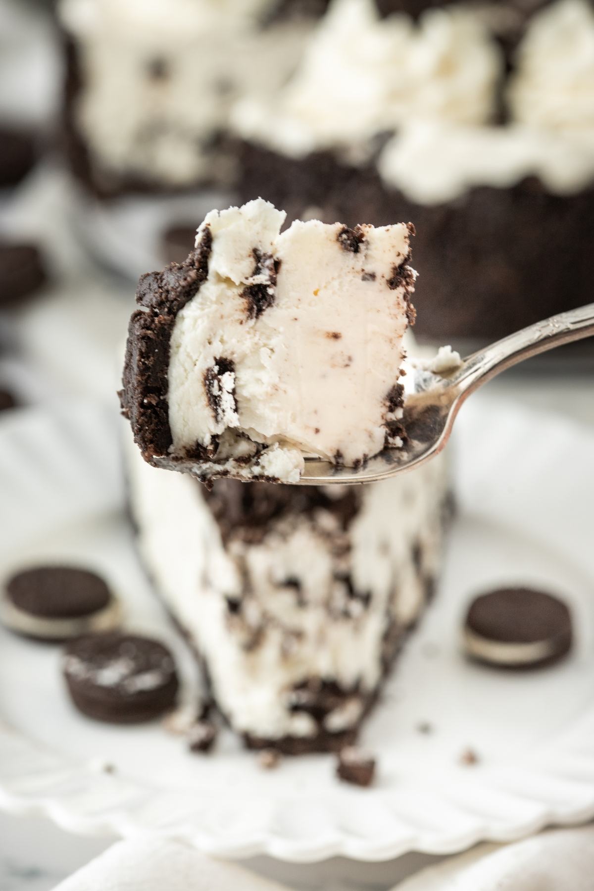 A fork holds a bite of cookies and cream cheesecake above a slice, with mini cookies on the plate.
