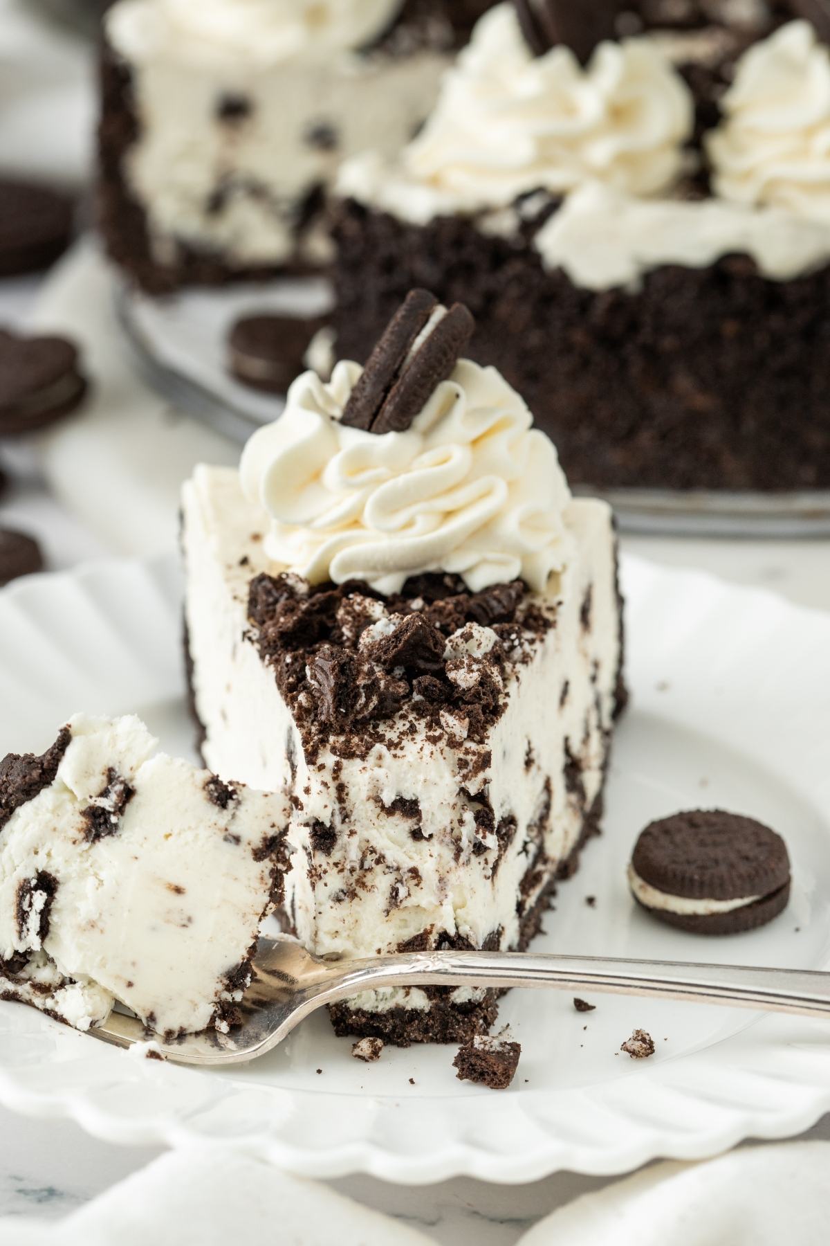 A slice of cookies and cream cheesecake topped with whipped cream and cookie pieces on a white plate.