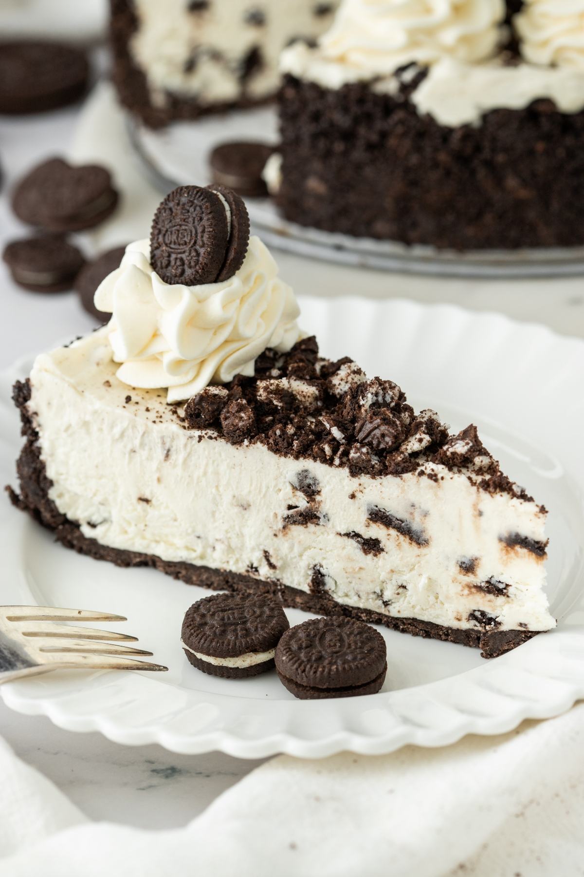 A slice of Oreo cheesecake on a plate, topped with whipped cream and mini Oreos, with more Oreos on the side.
