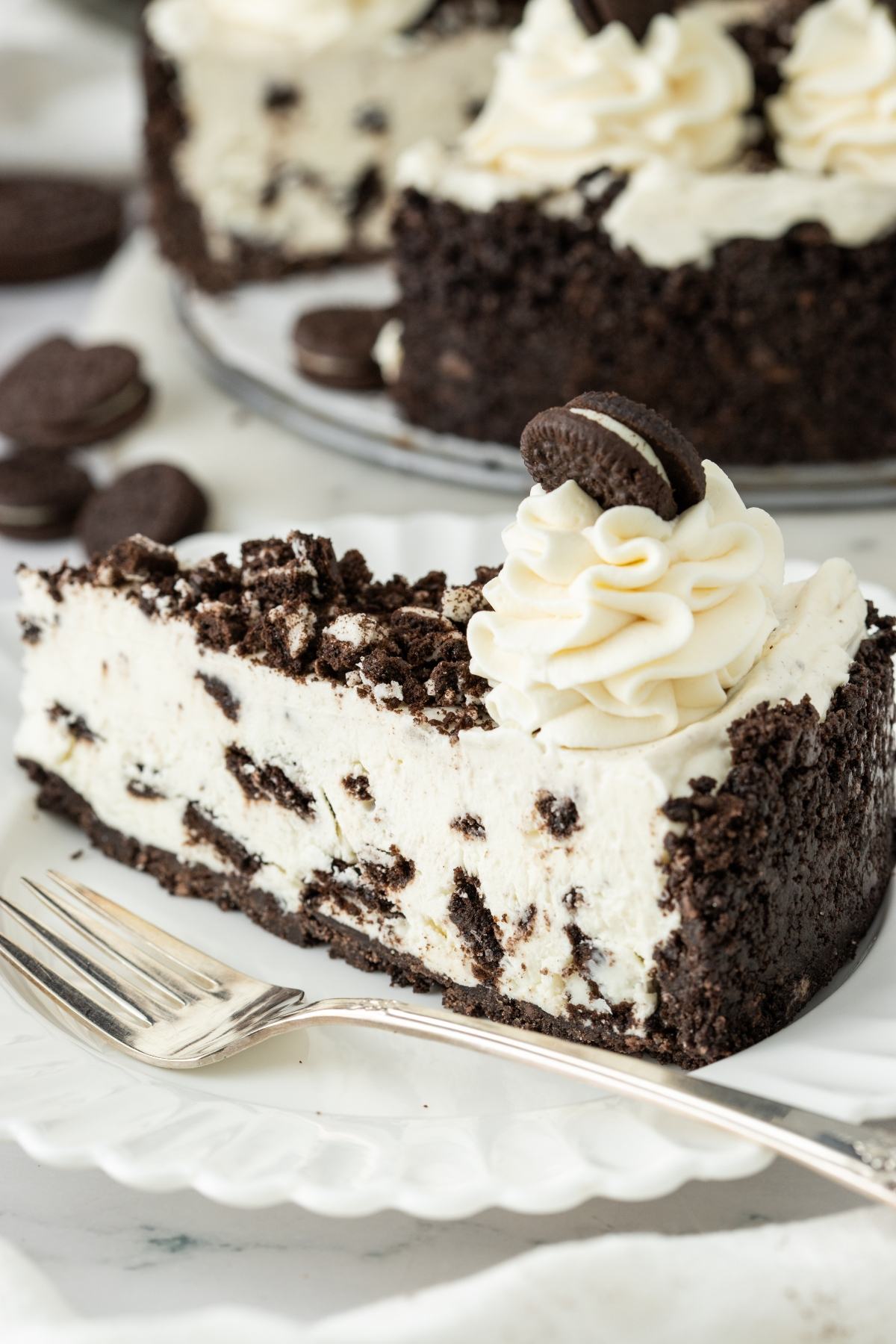 A slice of cookies and cream cheesecake with whipped cream and a mini cookie on top, on a white plate.