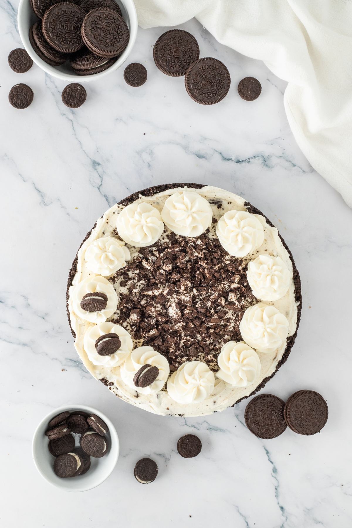 A cookies and cream cake topped with whipped cream swirls and chocolate sandwich cookies on a marble surface.
