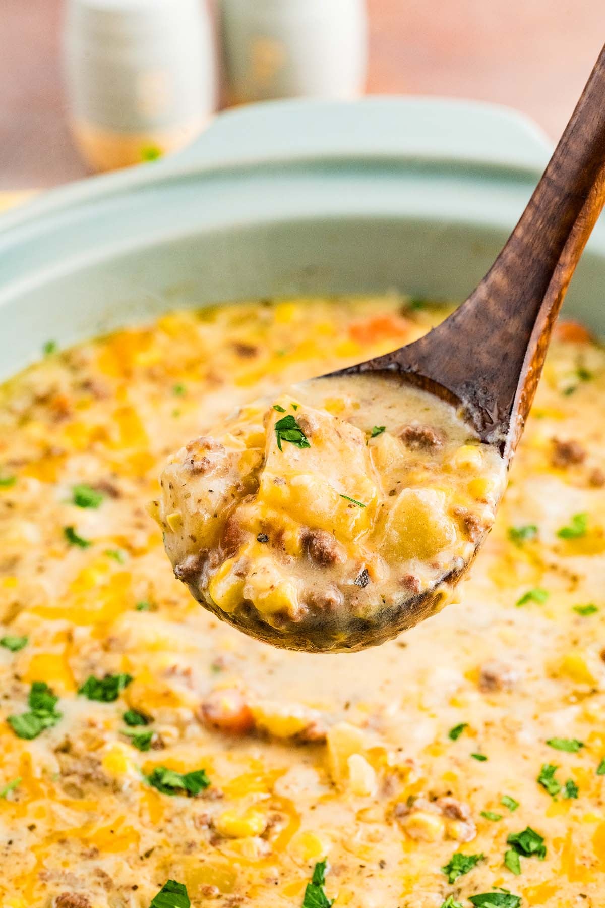 A wooden ladle holds a serving of Slow Cooker Cheeseburger Soup, filled with cheesy goodness, ground beef, potatoes, and herbs over a pot.