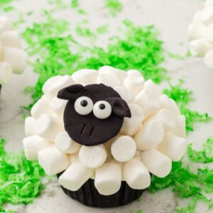 Sheep Cupcakes decorated with marshmallows and black fondant sit on green shredded coconut grass, creating an adorable farmyard treat.
