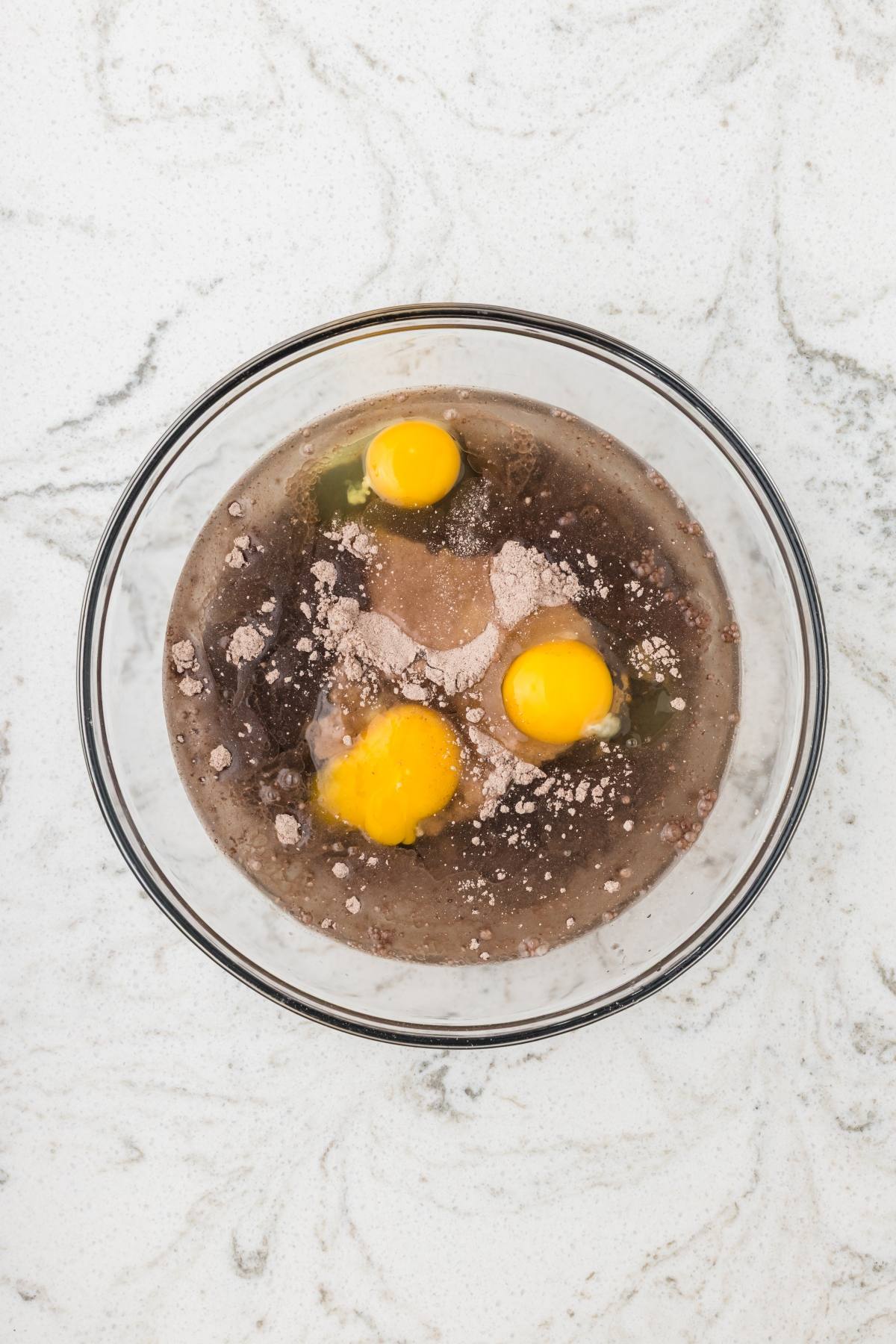 A glass bowl with three eggs, cocoa powder, and liquid ingredients on a white marble surface.