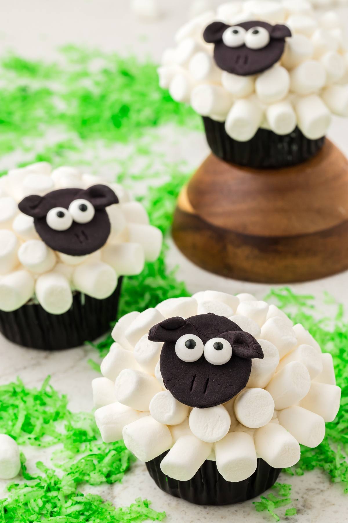 Cupcakes decorated as sheep with marshmallow wool and black fondant faces, on a white surface with green sprinkles.