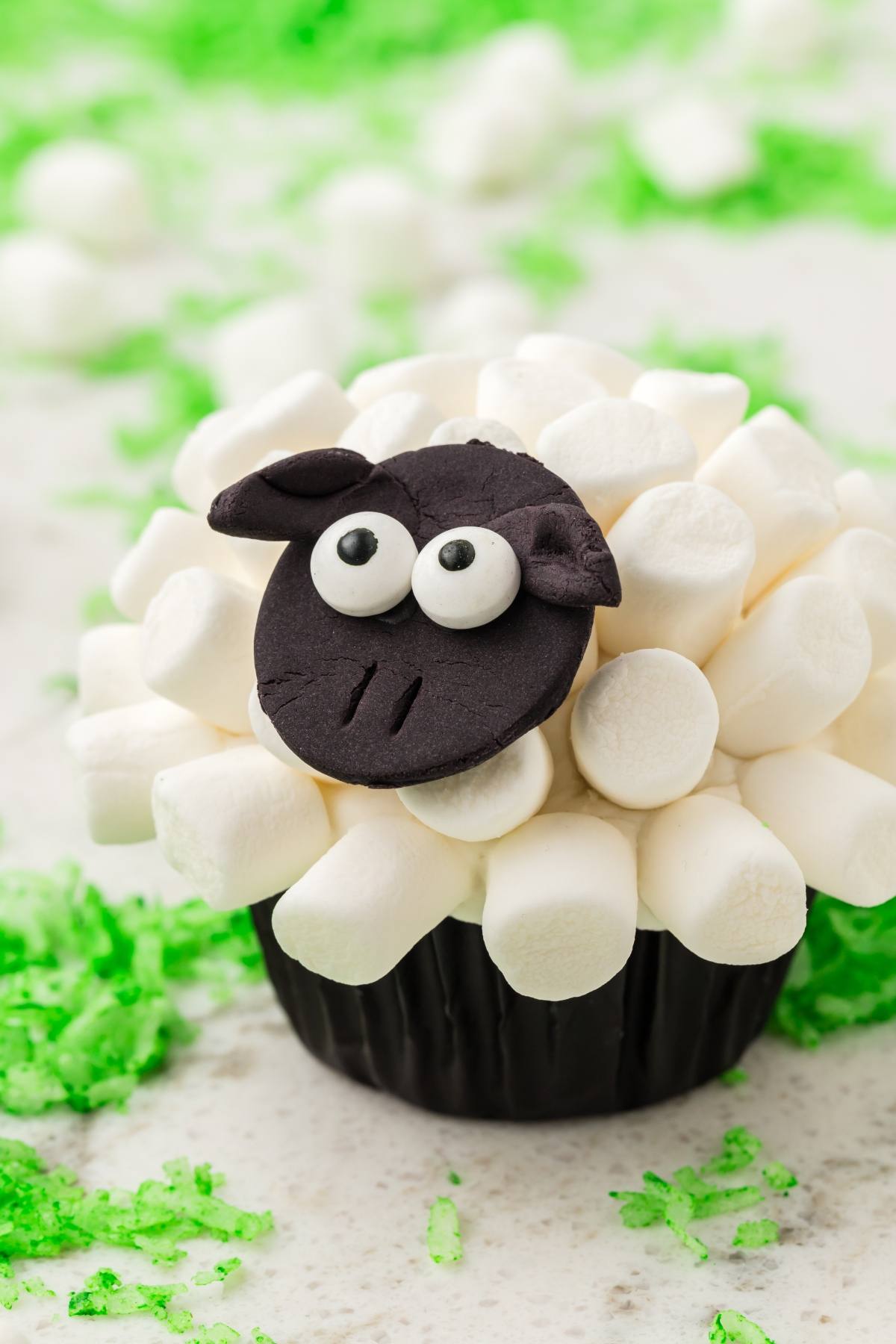 A cupcake decorated with mini marshmallows and candy to look like a sheep, on a light surface with green sprinkles.