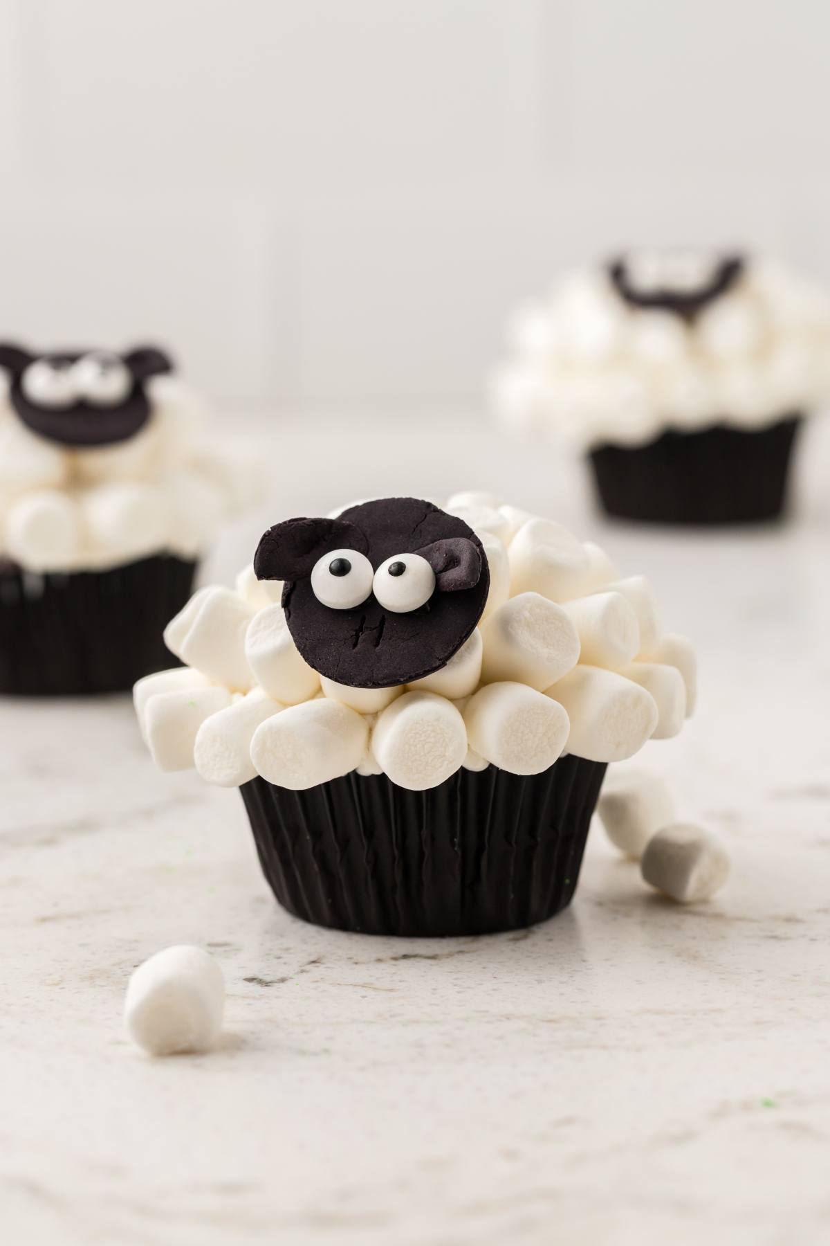 Chocolate cupcake decorated with marshmallows and a sheep face, with more sheep cupcakes in the background.