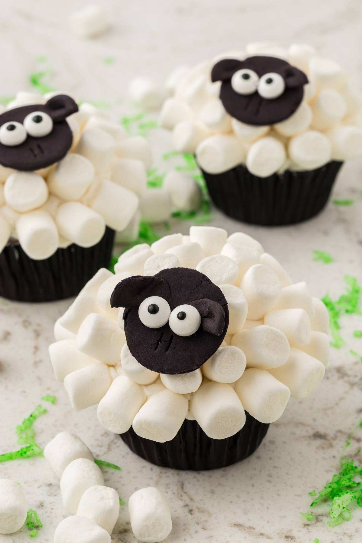 Cupcakes decorated with mini marshmallows and fondant sheep faces, arranged on a white surface.