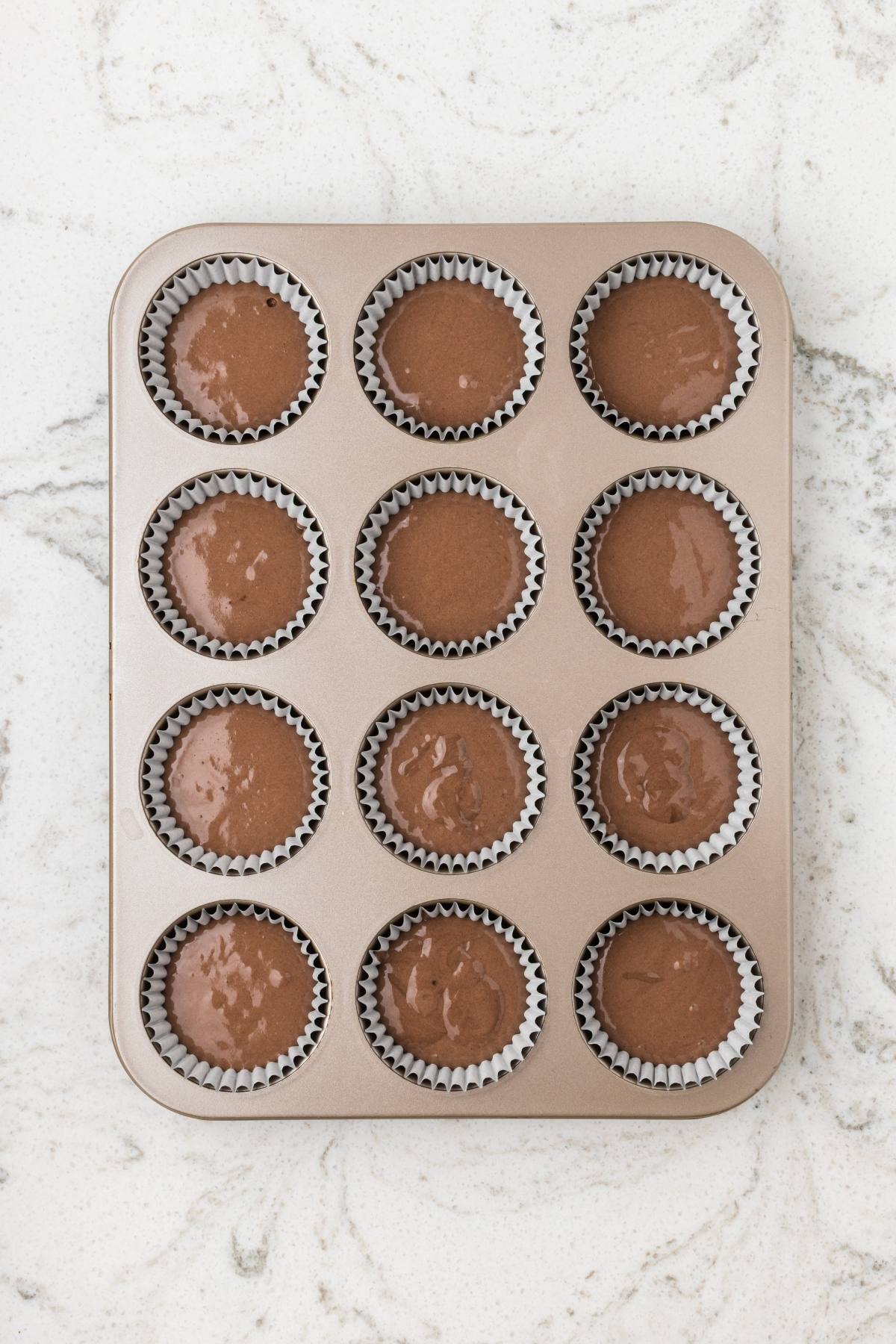 A muffin tin filled with chocolate batter in cupcake liners, sitting on a marble countertop.