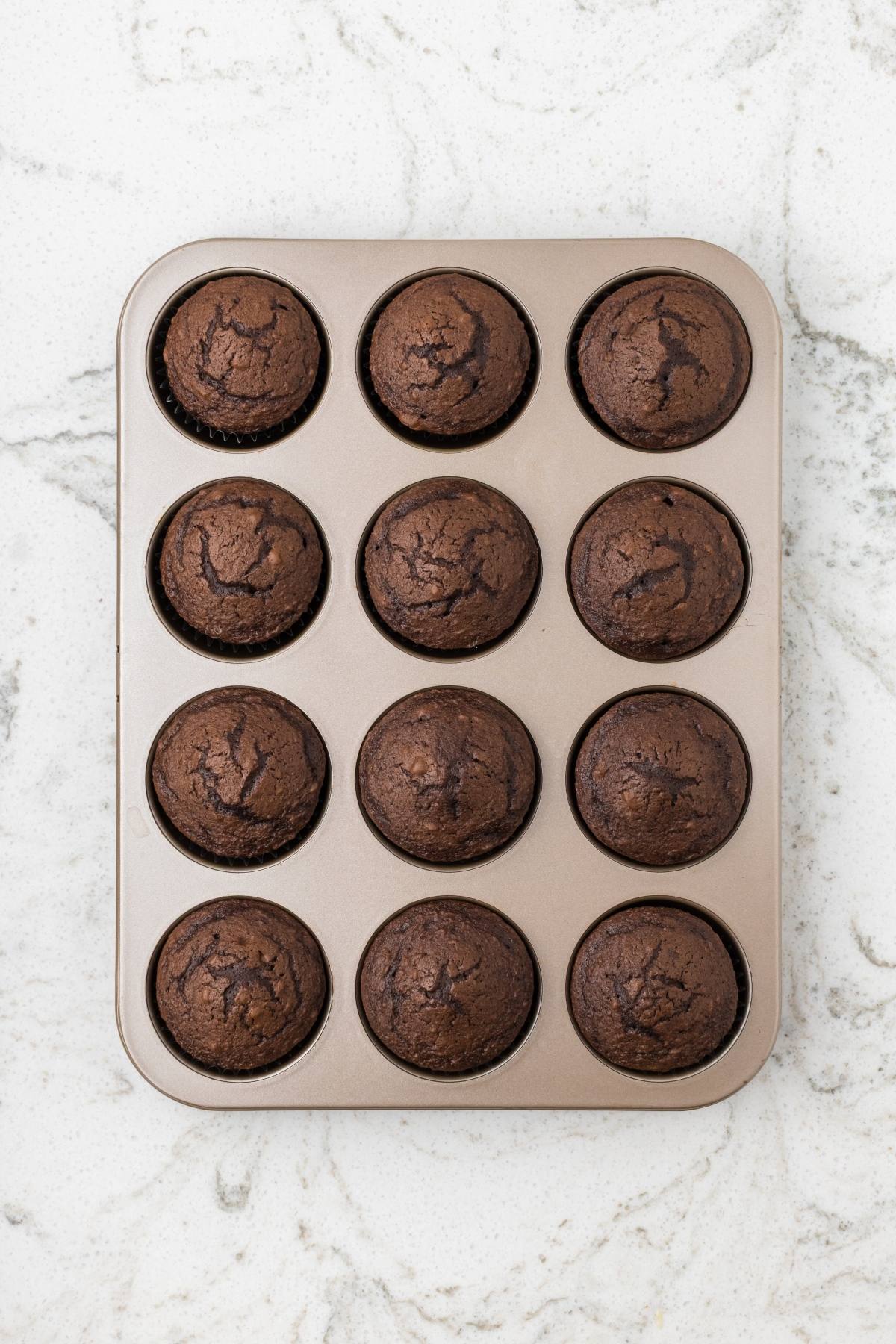 A muffin tin with twelve freshly baked chocolate muffins on a white marble countertop.