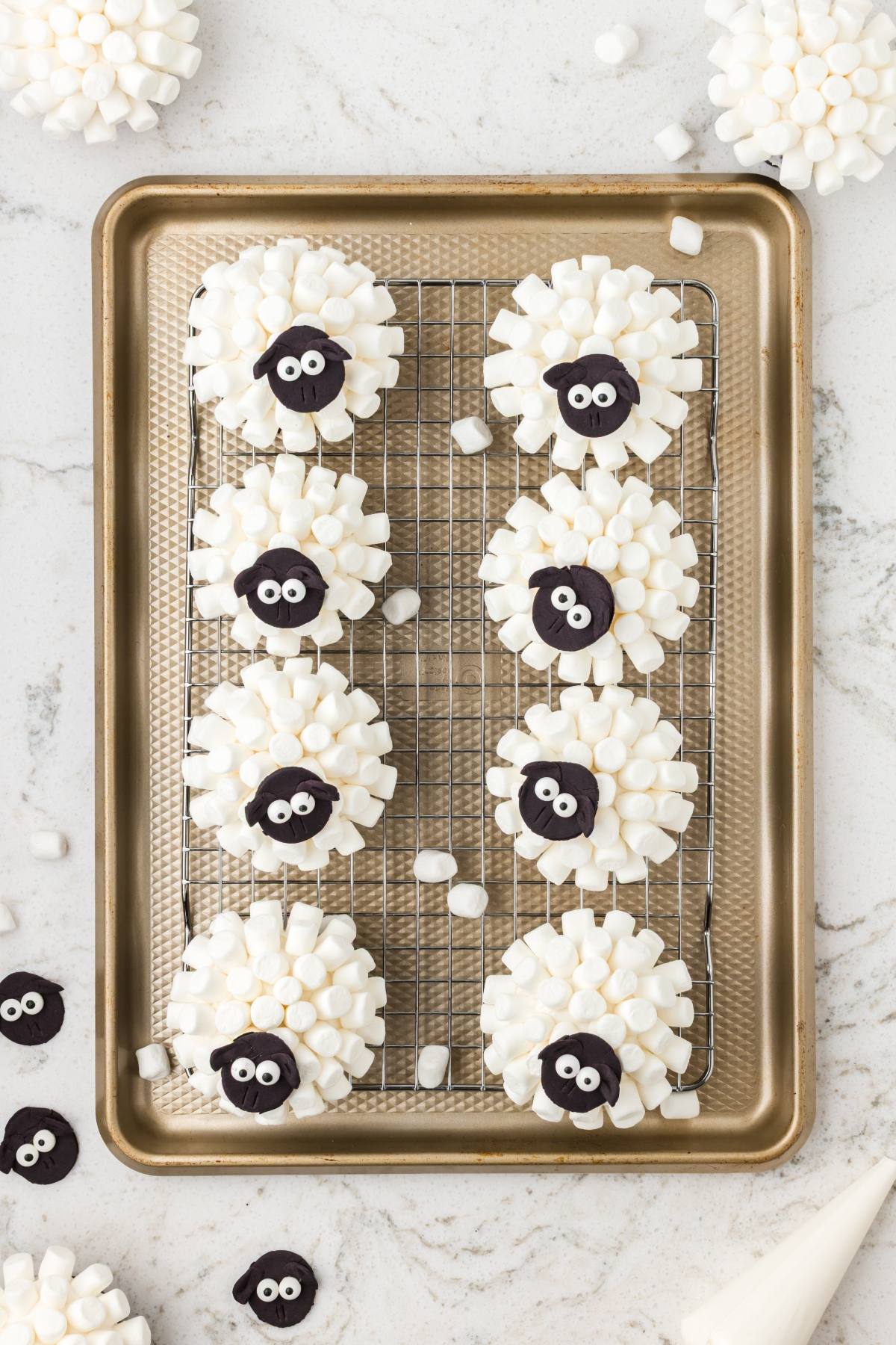 Eight sheep-themed cupcakes with white mini marshmallows and black candy faces on a wire rack and baking sheet.
