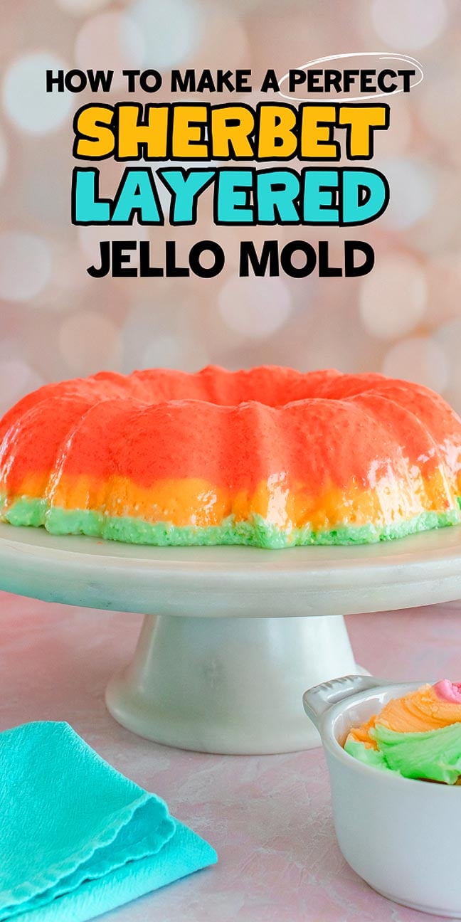 A vibrant Sherbet Layered Jello Mold sits on a cake stand, accented by a blue napkin and a pastel ice cream scoop nearby.