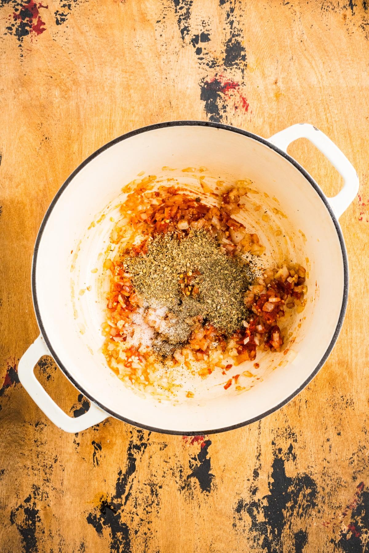 White pot with saut&eacute;ed onions, seasonings, and herbs on a rustic wooden surface.
