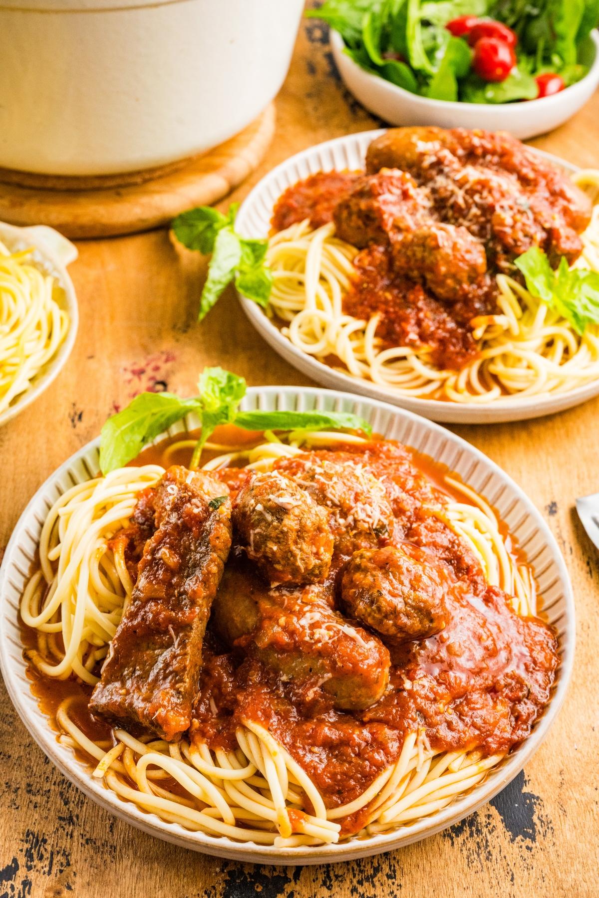Two plates of spaghetti topped with meatballs, sausage, red sauce, and garnished with basil. Salad in the background.