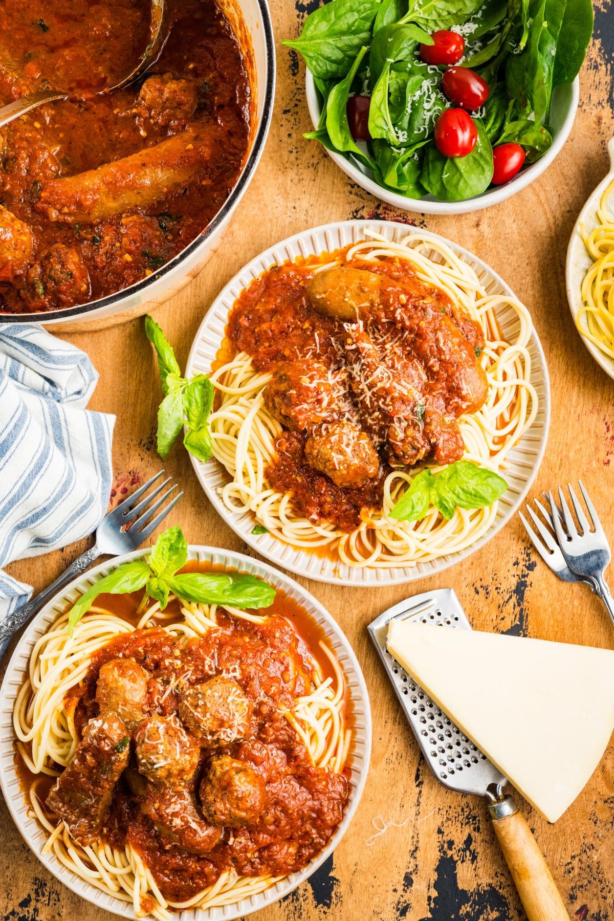 Plates of spaghetti with meatballs in tomato sauce, a bowl of salad, and a block of cheese on a table.