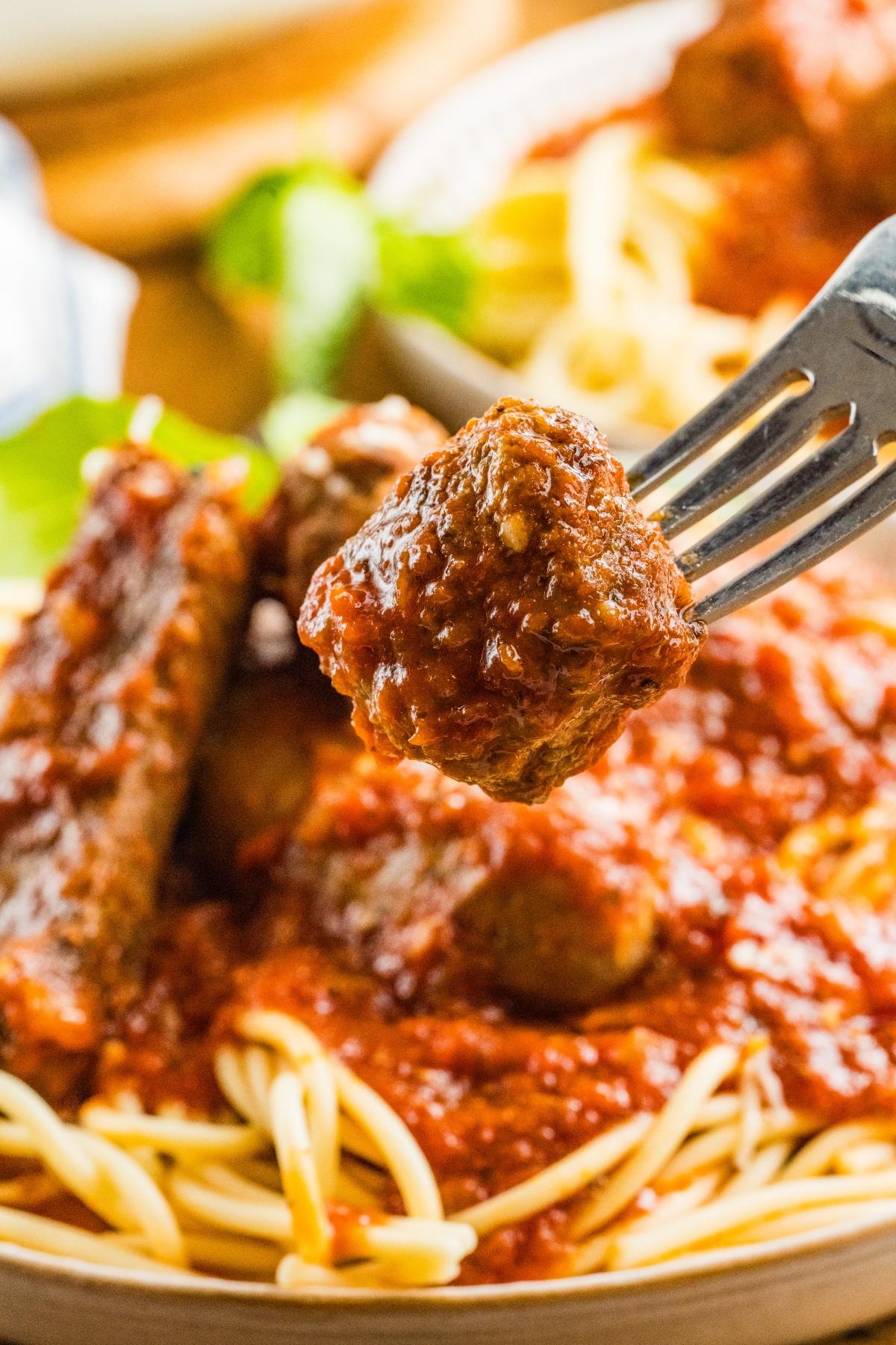 A fork holds a meatball above spaghetti topped with tomato sauce and more meatballs.