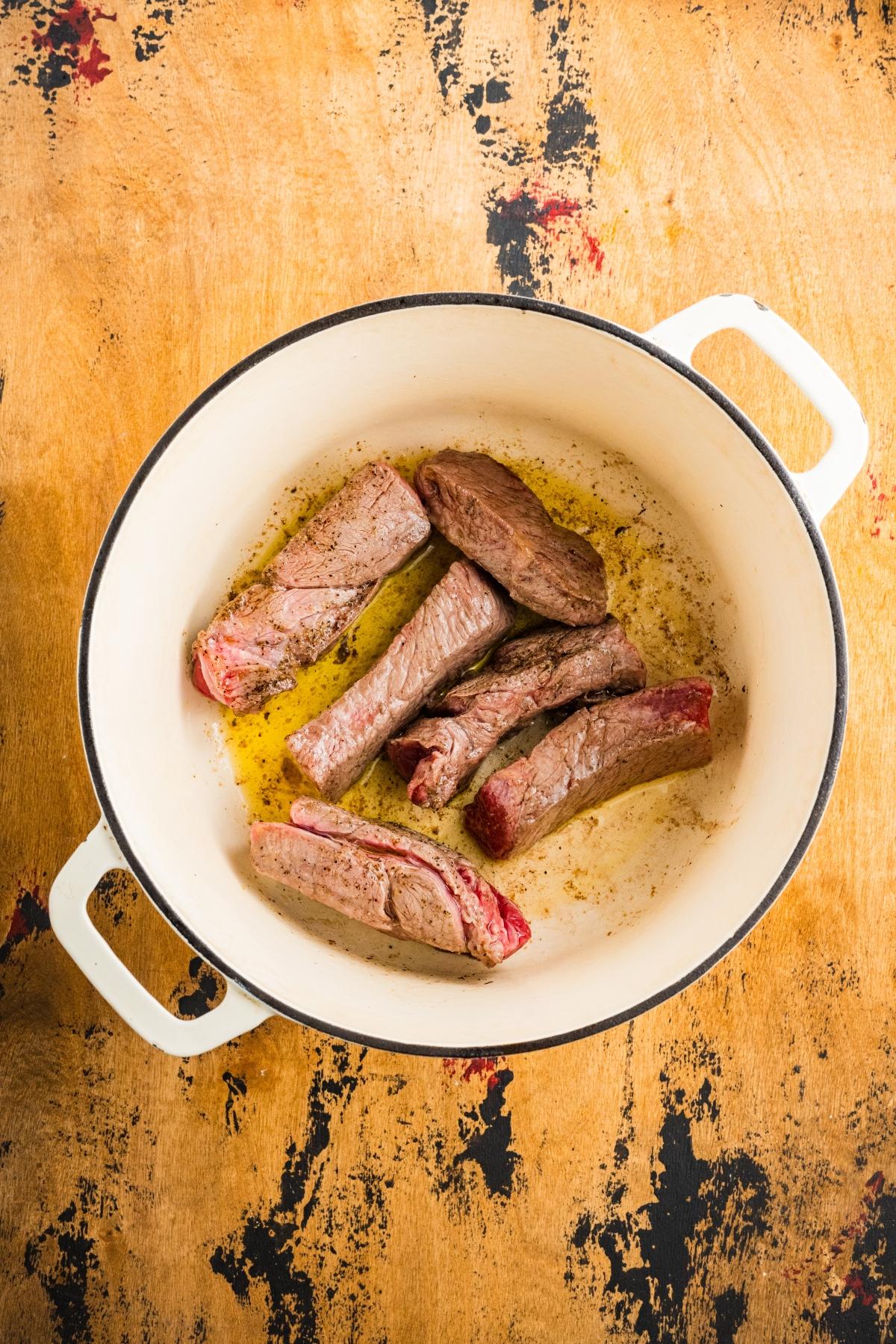 Five browned beef strips cooking in oil in a white pot on a rustic wooden surface.