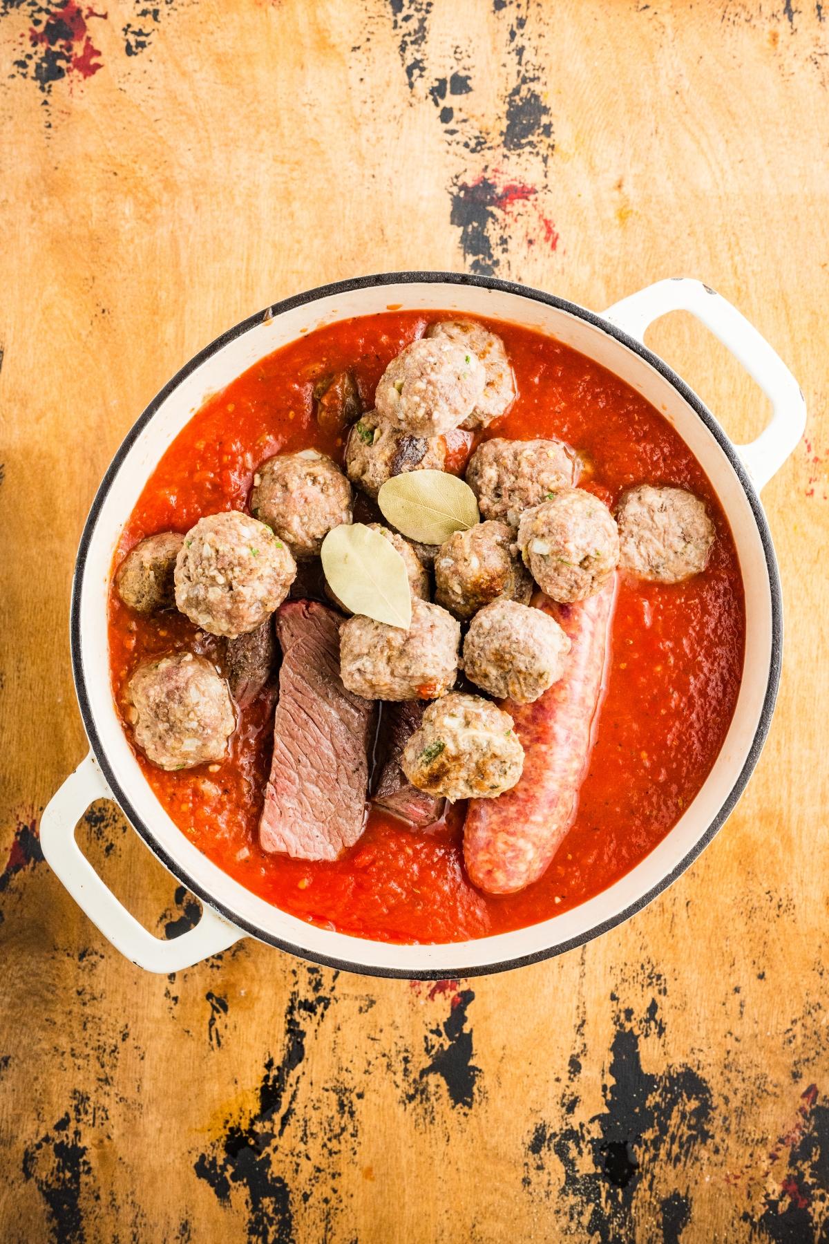 A pot of tomato sauce with meatballs, sausage, beef chunks, and bay leaves on a wooden surface.