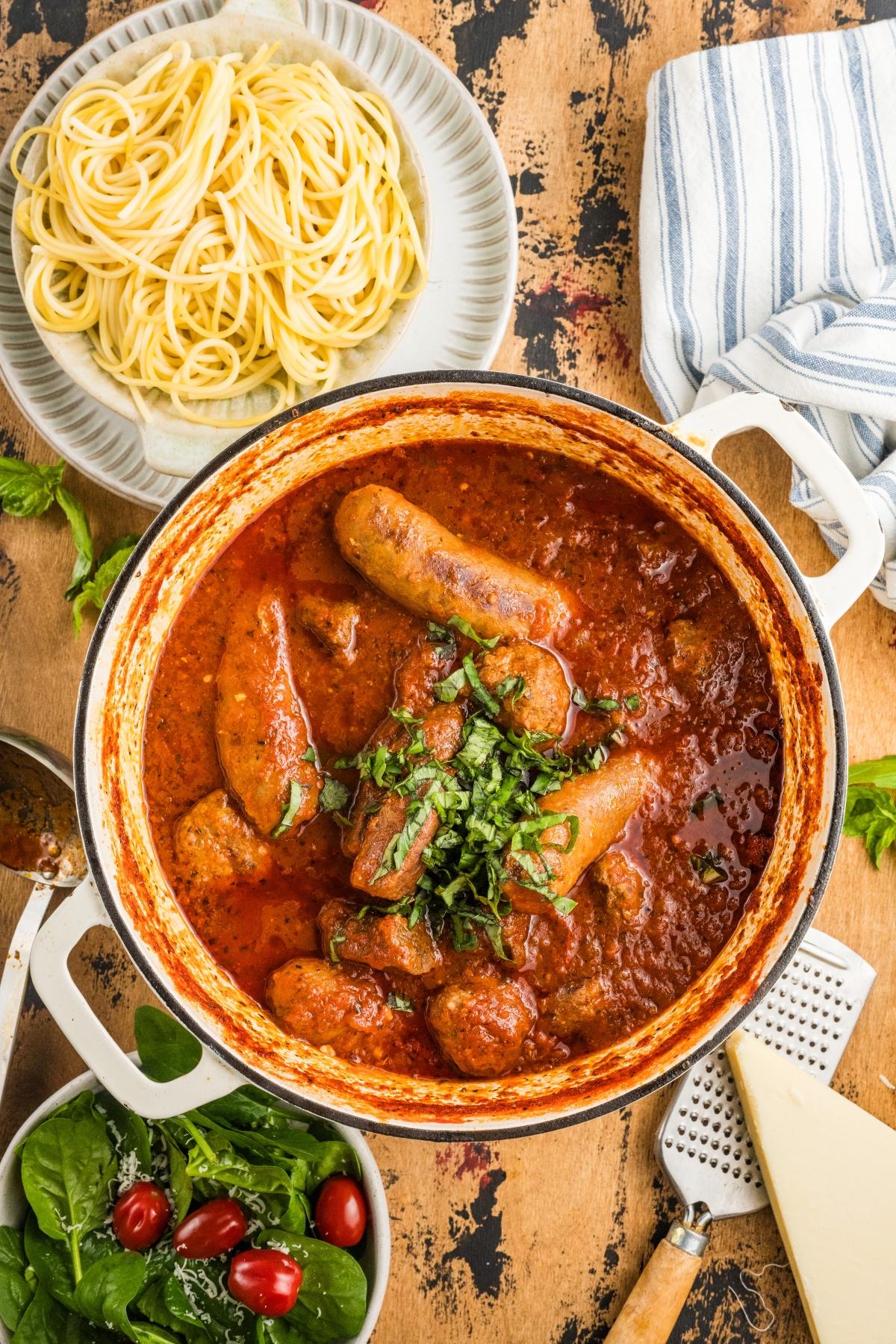 A pot of sausages in tomato sauce, a plate of spaghetti, and a salad with spinach and cherry tomatoes.