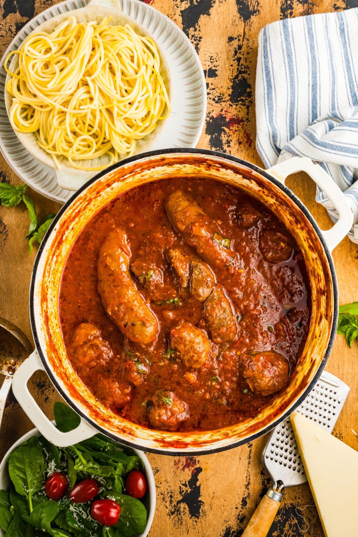 A pot of tomato sauce with sausages and meatballs, beside a plate of spaghetti and a salad.