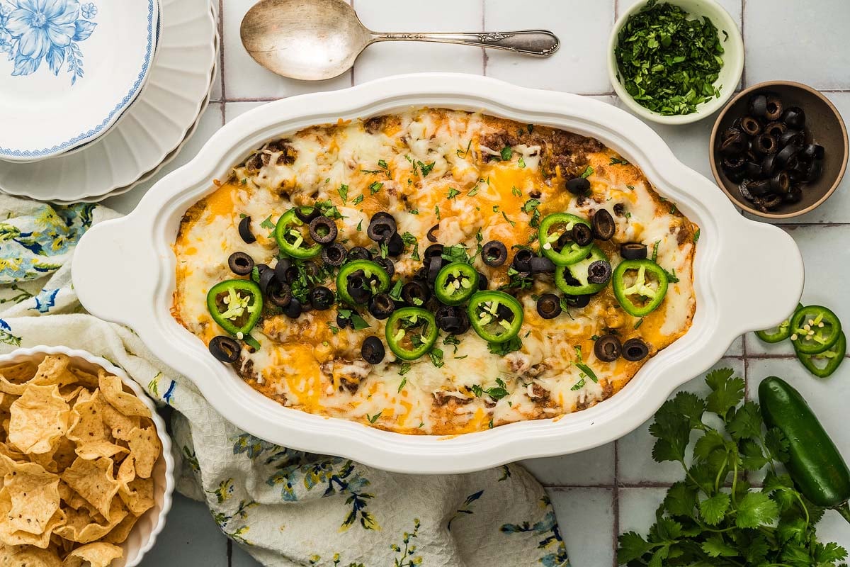 Texas Trash Dip baked casserole topped with melted cheese, black olives, and jalapeño slices, surrounded by chips and herbs.