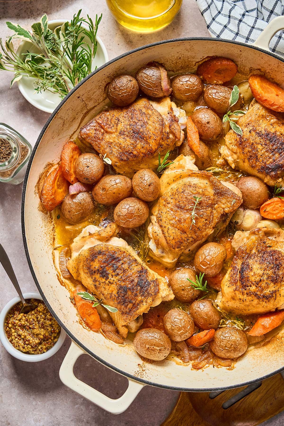 Beer Braised Chicken Thighs are roasted with baby potatoes and carrots in a large white pan, then garnished with fresh herbs.