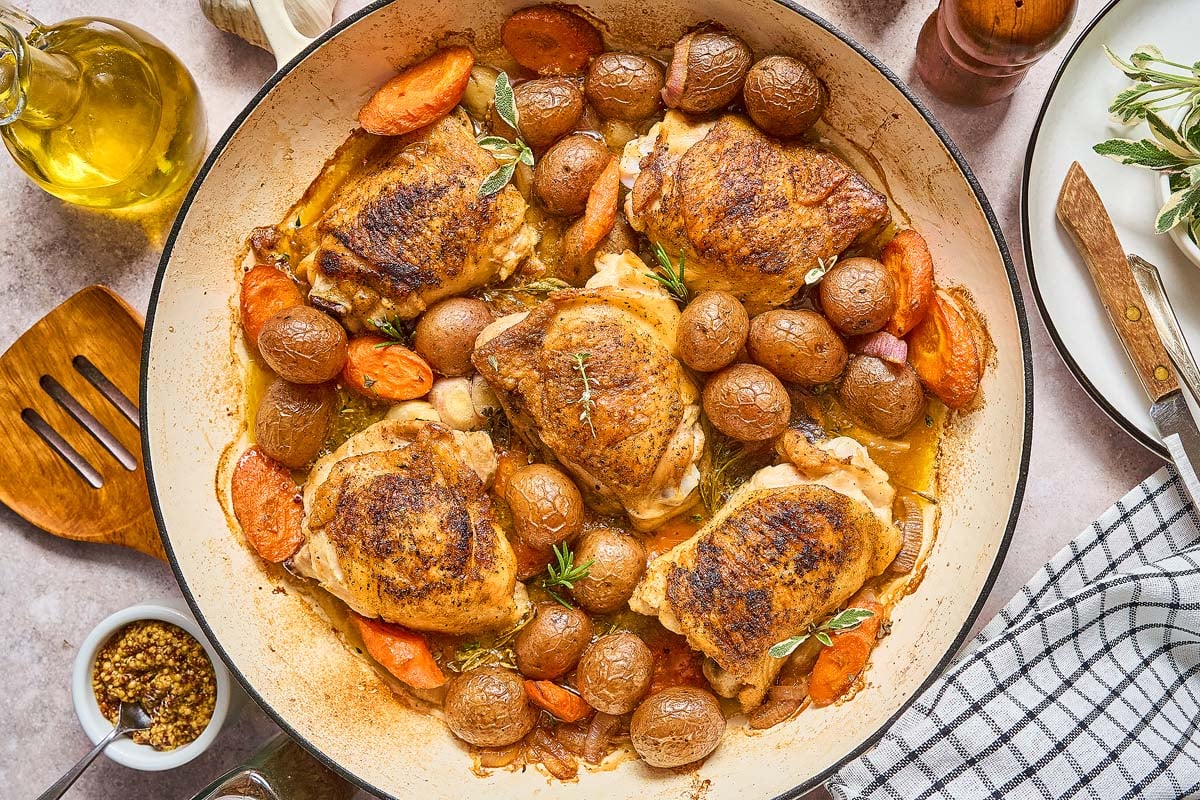 Beer Braised Chicken Thighs with baby potatoes and carrots in a large pan, surrounded by fresh herbs and kitchen utensils.